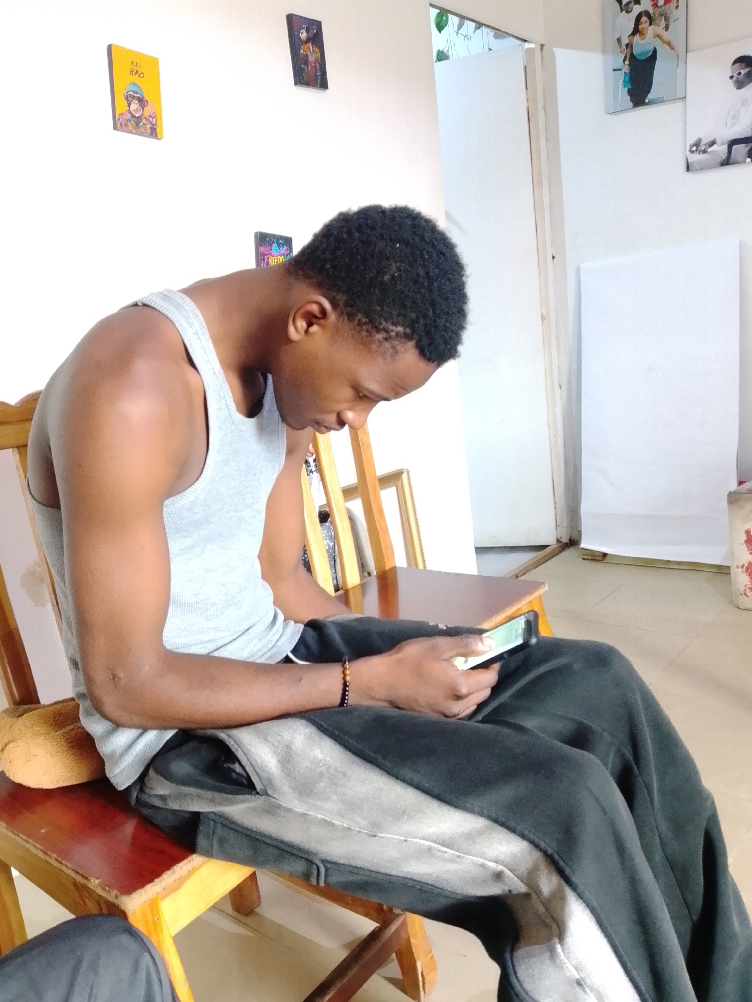 A young Black male wearing a grey tank top and dark grey sweatpants sits on a wooden chair in what appears to be a living room or common area. He is looking down at a smartphone in his hands, which is held horizontally. The man has short, dark curly hair and is wearing a beaded bracelet on his left wrist. To his right, a wooden framed picture hangs on the white wall. On the same wall, to the left, two framed pictures are also visible. The floor is tiled in a neutral color. The image is taken from a medium distance, capturing the man from the chest down. The lighting appears to be natural. The overall environment suggests a casual, indoor setting. There is no indication of the specific location within Jos, Nigeria, other than what can be inferred from the visual context.