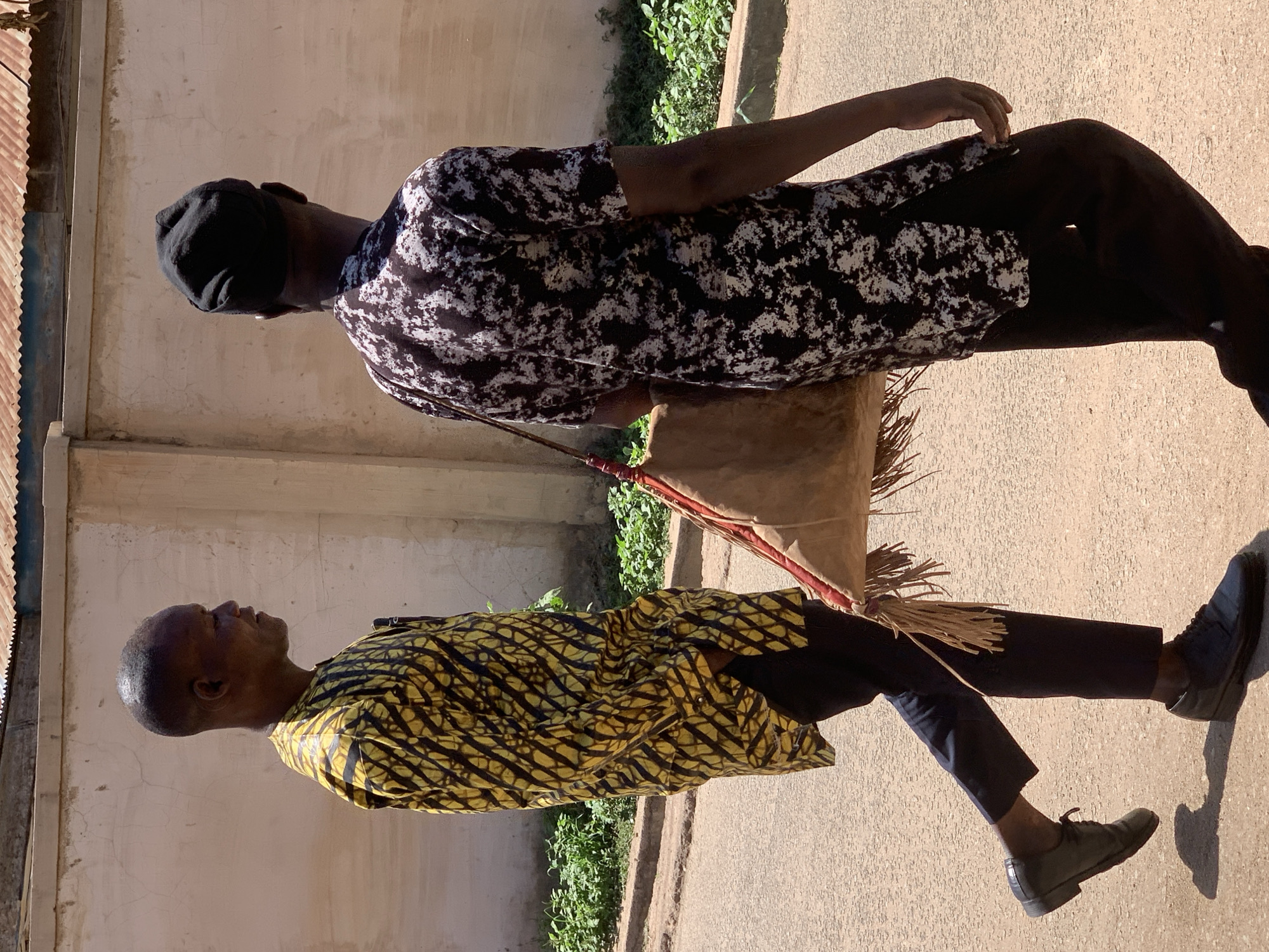 Two men are captured in an outdoor scene. The man on the left is wearing a yellow and black patterned shirt with dark trousers and dark shoes. The man on the right is wearing a white and dark patterned shirt, a black cap, dark trousers, and dark shoes. They are carrying a large, light brown sack-like object with fringe. The backdrop features a light-colored wall and some greenery. The location is inferred to be Jos, Nigeria.