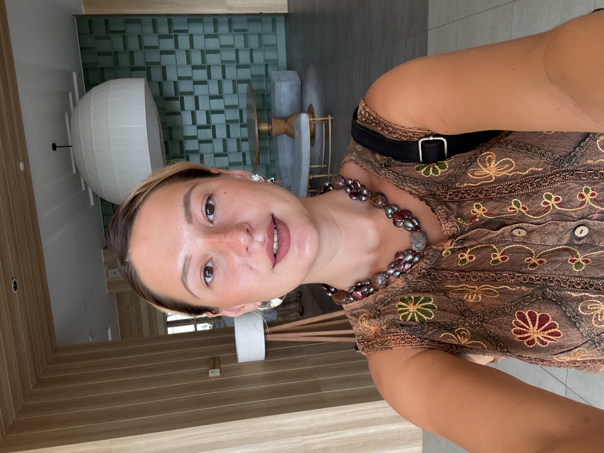 The image shows a woman taking a selfie in a modern indoor space. She is wearing a brown sleeveless top with floral embroidery and a beaded necklace. Her hair is pulled back, and she is wearing earrings. In the background, there are wooden paneled walls, a large white spherical light fixture, a green tiled wall, and some furniture, including a table and benches. The overall impression is that of a hotel lobby or a similar establishment. The image is undated and unlocated, and there is no information about the city of Da Nang, Vietnam.
