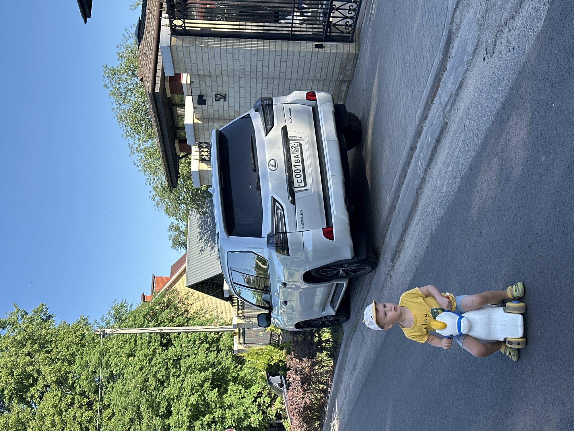 A young boy wearing a yellow t-shirt, blue shorts, and a white and yellow cap is riding a white and blue toy horse on a paved street. Behind him is a white Lexus LX parked on the side of the street. The license plate number visible on the Lexus is "C 001 BA 52 RUS". In the background are trees, a brick fence, and buildings. The environment suggests a residential area, and given the license plate, the location is within Nizhny Novgorod, Russia.