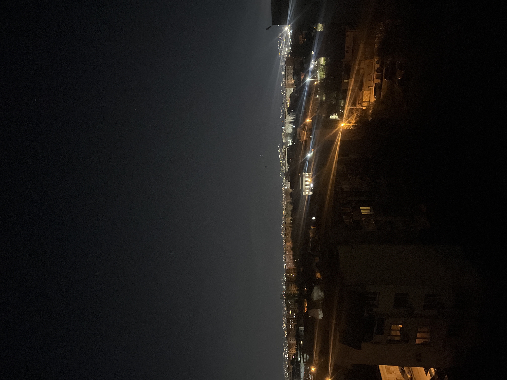 The image depicts a nighttime cityscape. The right side of the image shows buildings illuminated by streetlights and windows, while the left side is dark, possibly representing the night sky. The city is Sofia, Bulgaria. Buildings are densely packed. The scene is lit by artificial light from the city. There are no visible people.