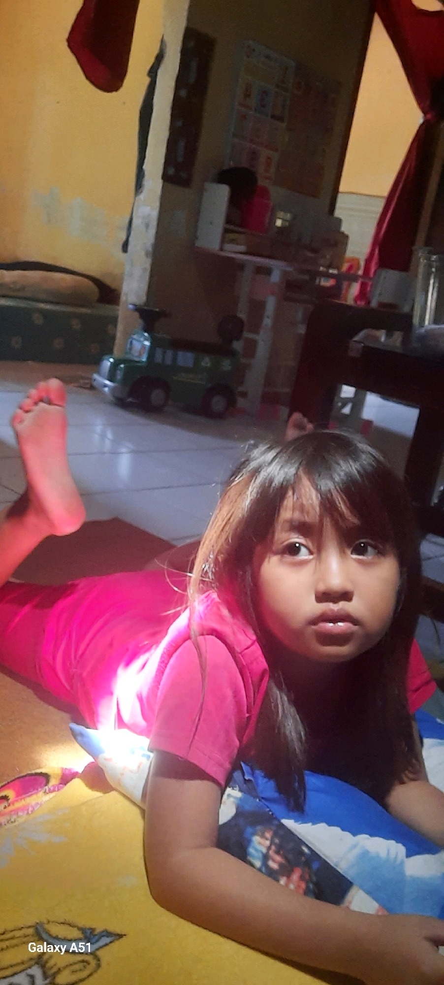 In Jatitengah, Indonesia, a young girl with dark hair and brown eyes is lying on her stomach on a yellow blanket. She is wearing a bright pink shirt and her left foot is visible, with its sole facing upwards. The girl is looking off to the right of the frame. In the background, a green toy truck is visible on a tiled floor. A table with some objects on it is also present, along with a red curtain. The lighting in the room appears to be artificial, casting a warm glow on the scene. The overall impression is of a casual, indoor setting.