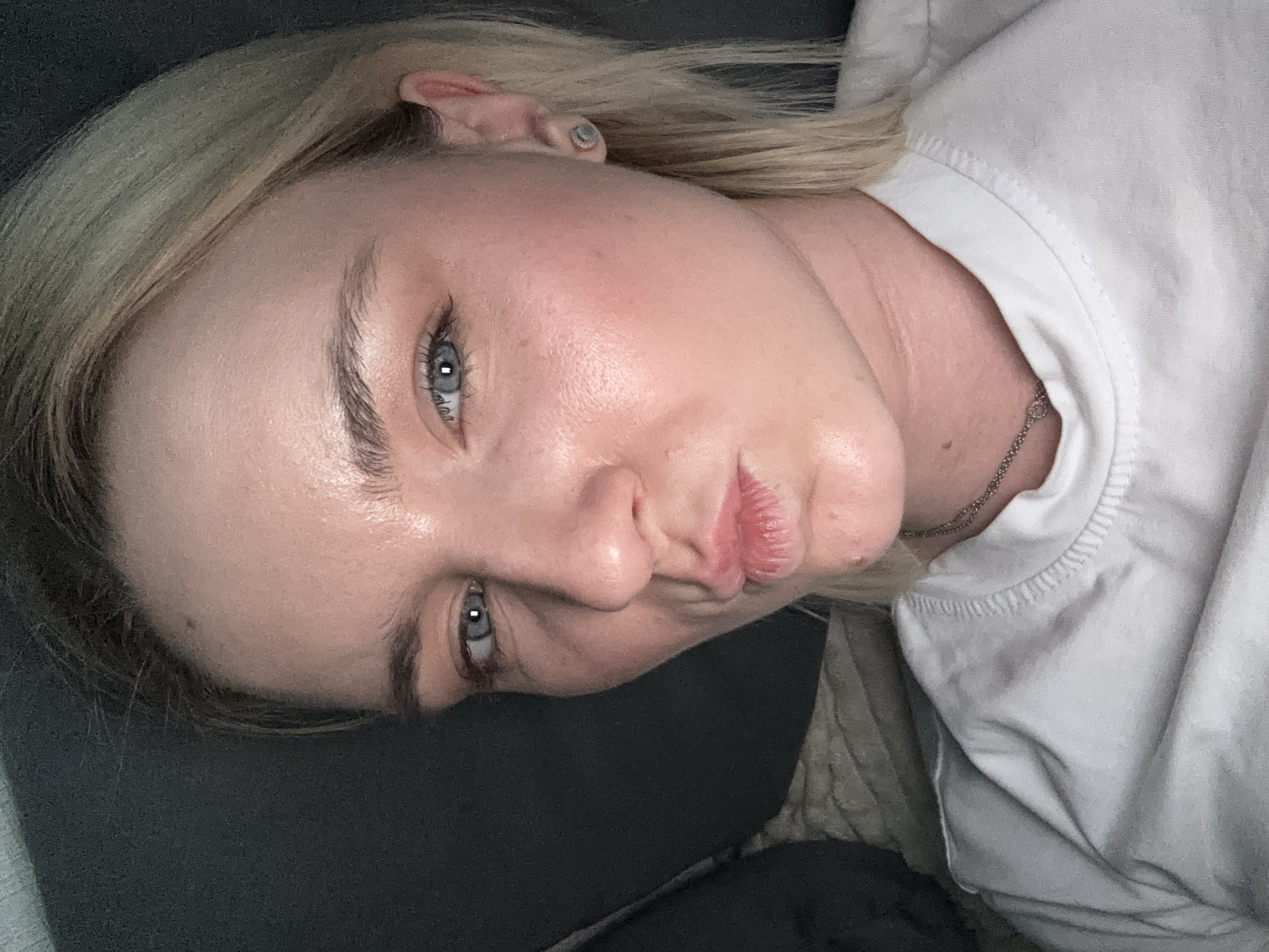 A young woman with blonde hair and blue eyes is lying down, angled towards the camera. She is wearing a white t-shirt and a silver chain necklace. Her lips are puckered in a slightly pouty expression. Her head rests against a dark, possibly gray, surface.
There is no indication of a location in Vsevolozhsk, Russia, or any other location. The image consists of a close-up portrait of the woman.