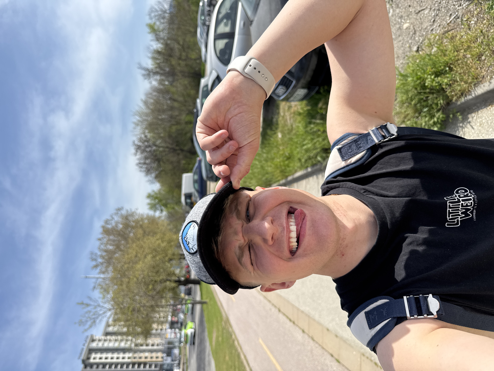 A young person poses playfully for a photo outdoors in an urban or suburban setting, likely Plovdiv, Bulgaria, given the context. The person is seen from the chest up, with their head tilted, looking directly at the camera. They are grinning widely with squinted eyes and their tongue playfully sticking out.
The individual is wearing a grey and black baseball cap with a light-colored circular logo on the front, which they are touching with their left hand. A light beige or white smartwatch with several holes in the strap is visible on their left wrist. They are dressed in a black t-shirt that prominently displays white text on the chest, reading "LITTLE WEIRD" in a stylized font, with smaller text below saying "EVERYBODY IS A LITTLE WEIRD". A dark blue and white strap, possibly from a backpack or sling bag, with a silver-colored buckle is visible over their right shoulder.
The background shows a paved area, suggesting a street or parking lot, with several cars parked. Beyond the cars, there are trees with budding leaves and distant multi-story buildings under a bright blue sky with some scattered white clouds. The clear skies and strong shadows indicate it is daytime, likely a sunny day. The overall impression is one of lightheartedness and joy.