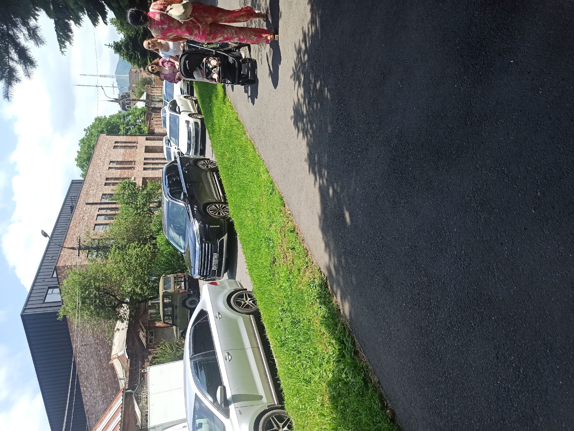 In Atabe, Georgia, a paved walkway runs parallel to a street lined with parked cars and a grassy verge. On the walkway, a person pushes a stroller with a baby. Further down the street, several cars are parked, including a silver sedan and a black SUV. Behind the cars, a modern brick building with a dark metal roof stands against a backdrop of trees and a clear sky. The scene depicts a casual urban environment on a sunny day.