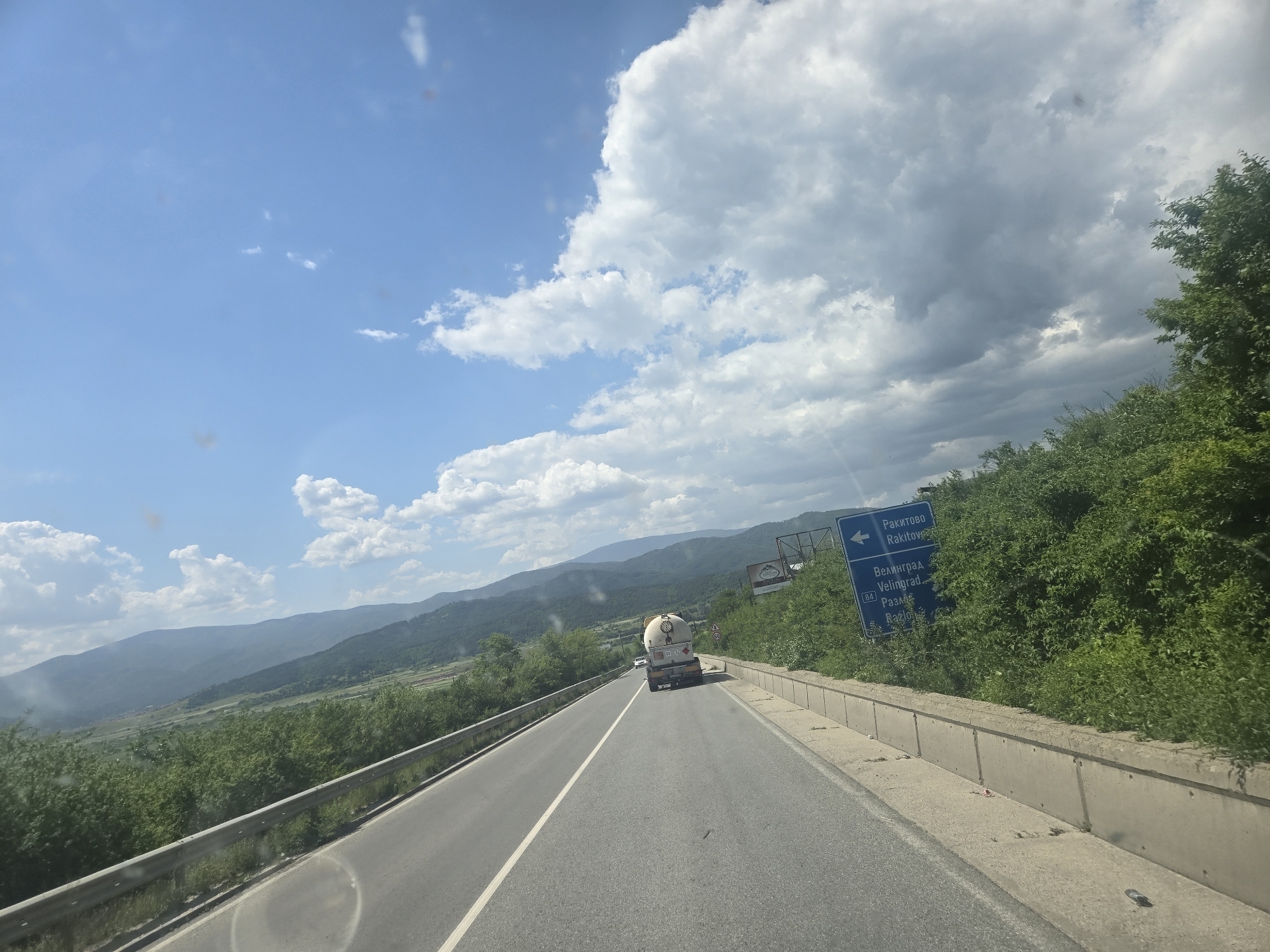 The image depicts a road in a mountainous region, potentially a highway. A truck, possibly a tanker, is traveling in the same direction as the viewer. The sky is partly cloudy with patches of blue. A road sign indicates directions to "Ракитово Rakitovo" and "Велинград Velingrad Разлог Razlog" indicating that the location is likely in Bulgaria. Dense green vegetation covers the slopes adjacent to the road. There are mountains visible in the distance.