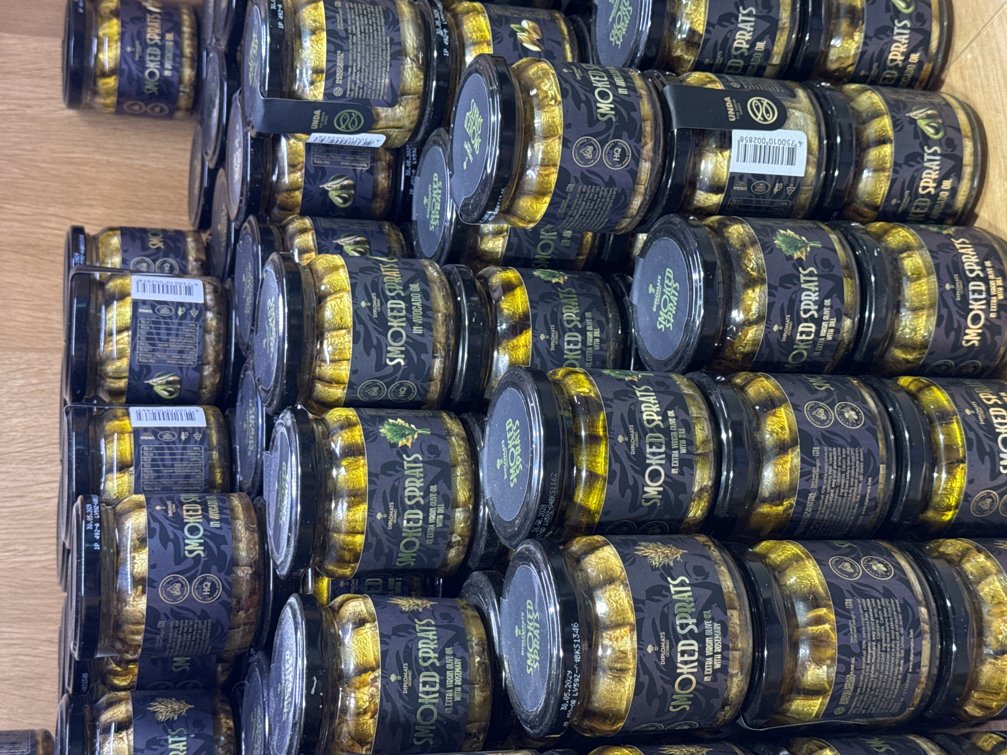 The image shows a close-up of a stack of glass jars filled with smoked sprats preserved in oil. The jars are arranged in a haphazard manner on a wooden surface. The jars have black labels with the "Diplomats Gourmet" brand name. Some labels indicate the sprats are in extra virgin olive oil with dill or rosemary, while others say they are in avocado oil. A label on a jar says the product is made in Riga, Latvia, by SIA Unda.