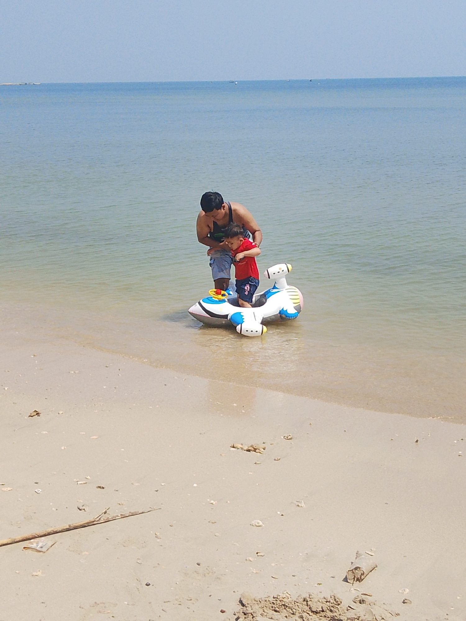 In the city of Pejagan, Indonesia, a man and a young child are standing in shallow water near the shore. The man, wearing a dark tank top and blue shorts, is holding the child. The child, wearing a red t-shirt and blue shorts, is standing on an inflatable toy that resembles a white airplane with blue accents and a yellow steering wheel. The water is calm and clear, with the sandy beach in the foreground. The sky is a pale blue.