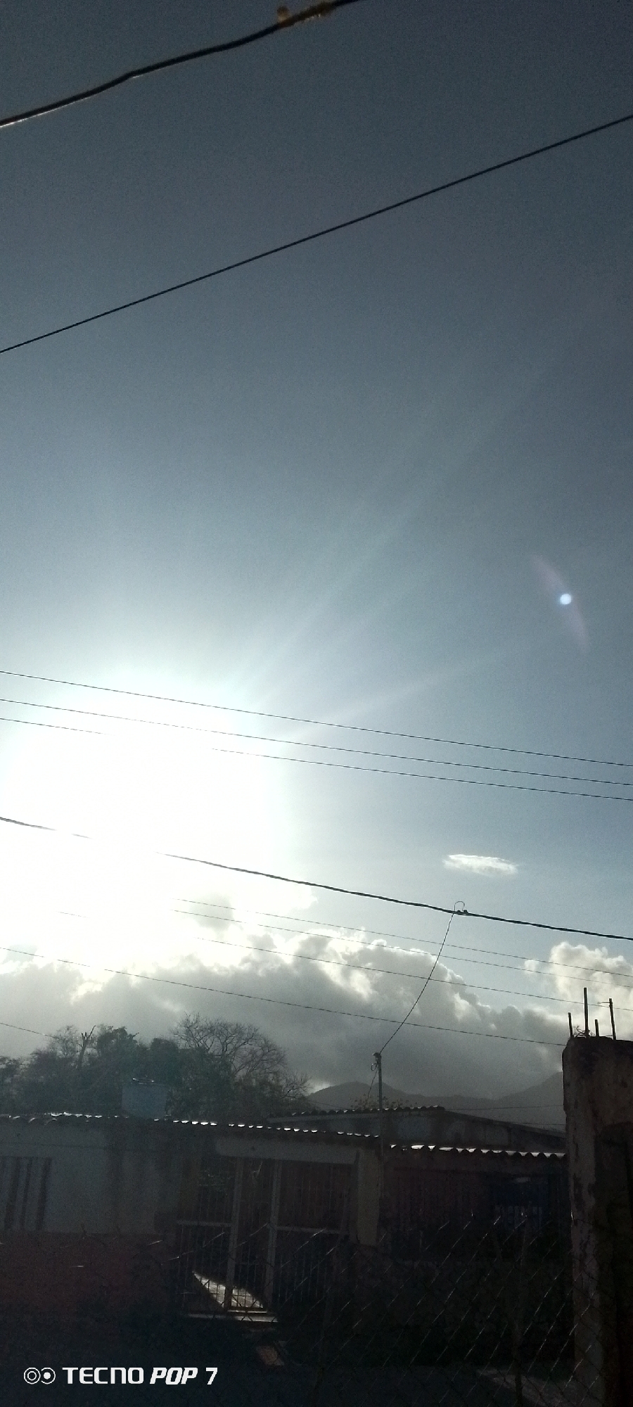The image shows a bright, slightly overcast sky with the sun obscured by clouds. Power lines run across the frame. In the lower portion, there are buildings and trees. A fence is visible in the foreground. The location appears to be a residential area, possibly in Altagracia, Venezuela.