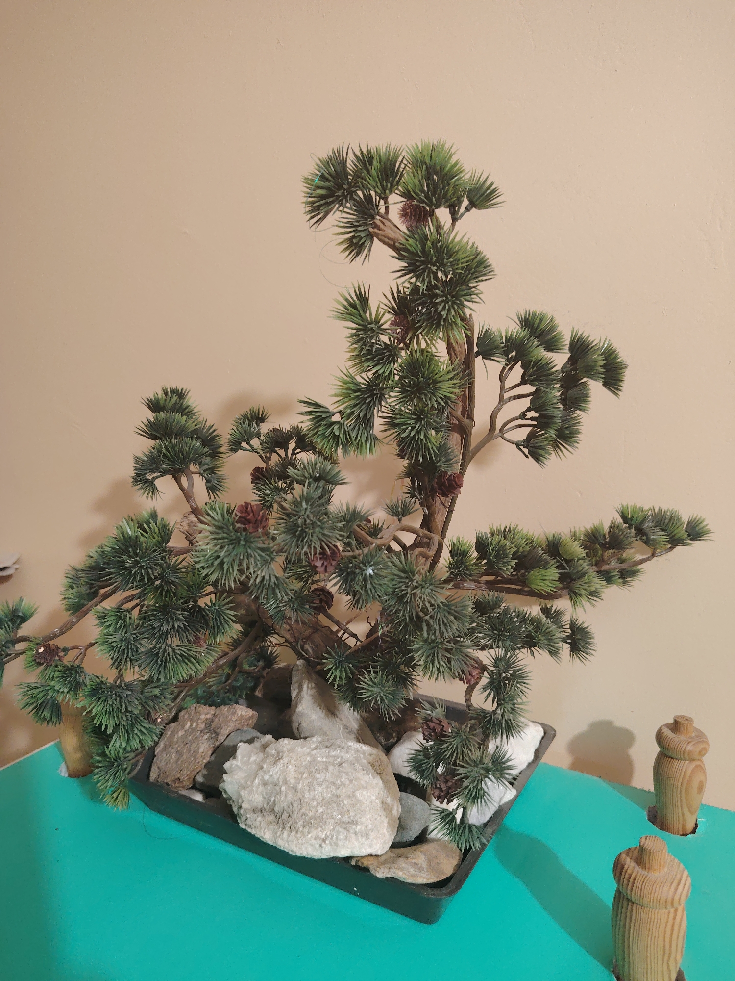 A faux pine bonsai tree with artificial pinecones sits in a dark rectangular planter filled with white decorative stones and larger rocks. The planter rests on a teal-colored surface. In the background, a beige wall is visible. To the right of the planter, two small, turned wooden decorative objects protrude from the teal surface. The lighting casts a soft shadow of the tree and the wooden objects onto the wall behind them.