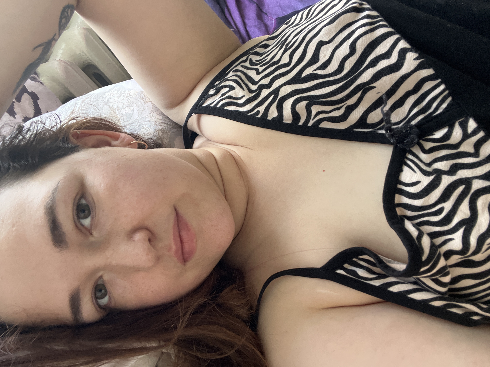 A woman with long auburn hair lies on a bed, looking at the camera. She is wearing a black and white zebra-print camisole. The background consists of a pillow and bedding. The image appears to be a selfie taken indoors. There is no information in the image to suggest a location in Chelyabinsk, Russia.