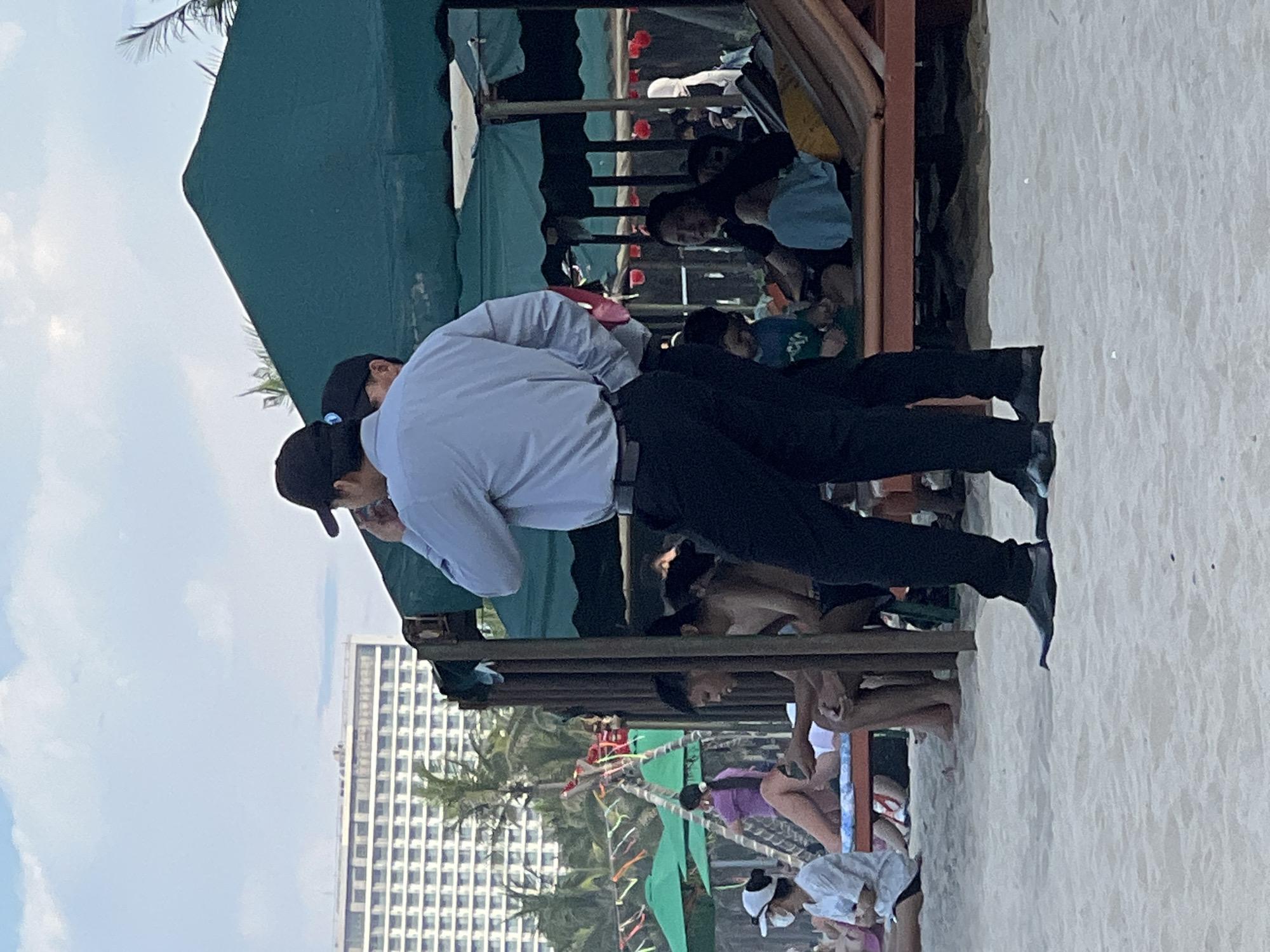 The image shows a beach scene in Da Nang, Vietnam. In the foreground, two men in dark pants and light-colored shirts, wearing baseball caps, stand facing away from the camera. One man is holding a phone to his ear. Behind them, several people are seated under a large green canopy. Some are dressed in beachwear, while others are more casually dressed. In the background, a large, multi-story building with many windows is visible, along with palm trees and smaller green umbrellas, suggesting a resort or hotel area. The sky is cloudy. The sand on the beach appears light in color.