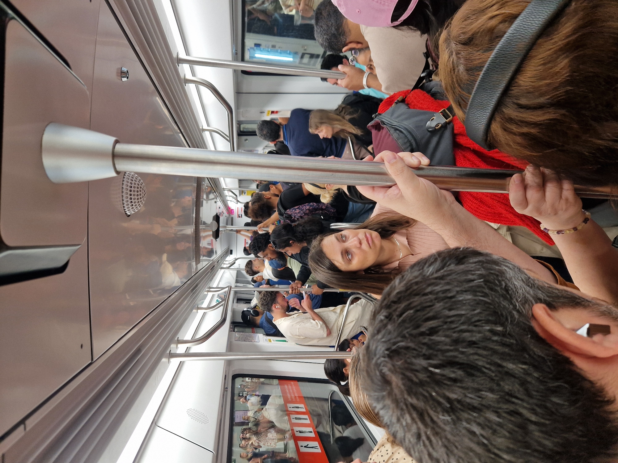 The image depicts the interior of a Barcelona Metro train carriage. Numerous people are visible, standing close together. Some are looking at their phones, while others are looking around. The train car features stainless steel handrails running along the ceiling for standing passengers to hold. The walls of the train are a light gray. There are windows with advertisements and safety information displayed on them. The overall atmosphere is crowded.