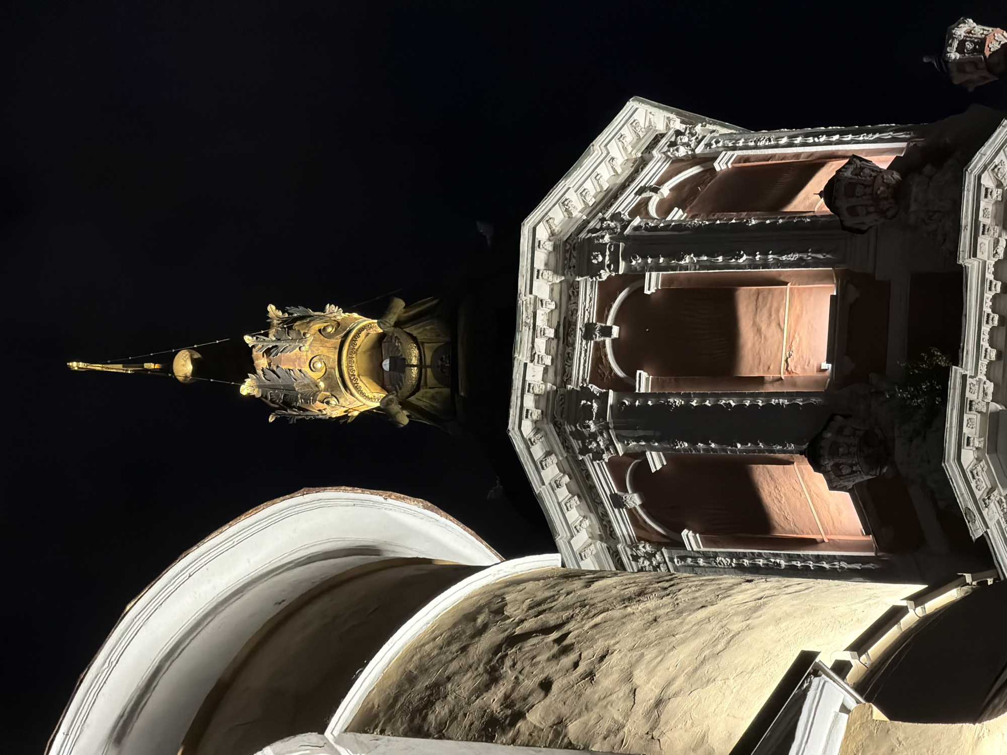 At night, a low-angle shot looks up at two distinct sections of the Cathedral of Christ the Savior in Moscow, Russia.
The lower foreground shows the textured surface of a large dome, illuminated with a warm light from below. Above it, the lower portion of the tiered section of the cathedral is partially visible, with ornately decorated arches and niches.
Further above and to the left, the ornate gold-plated top of a bell tower is lit against the dark night sky.