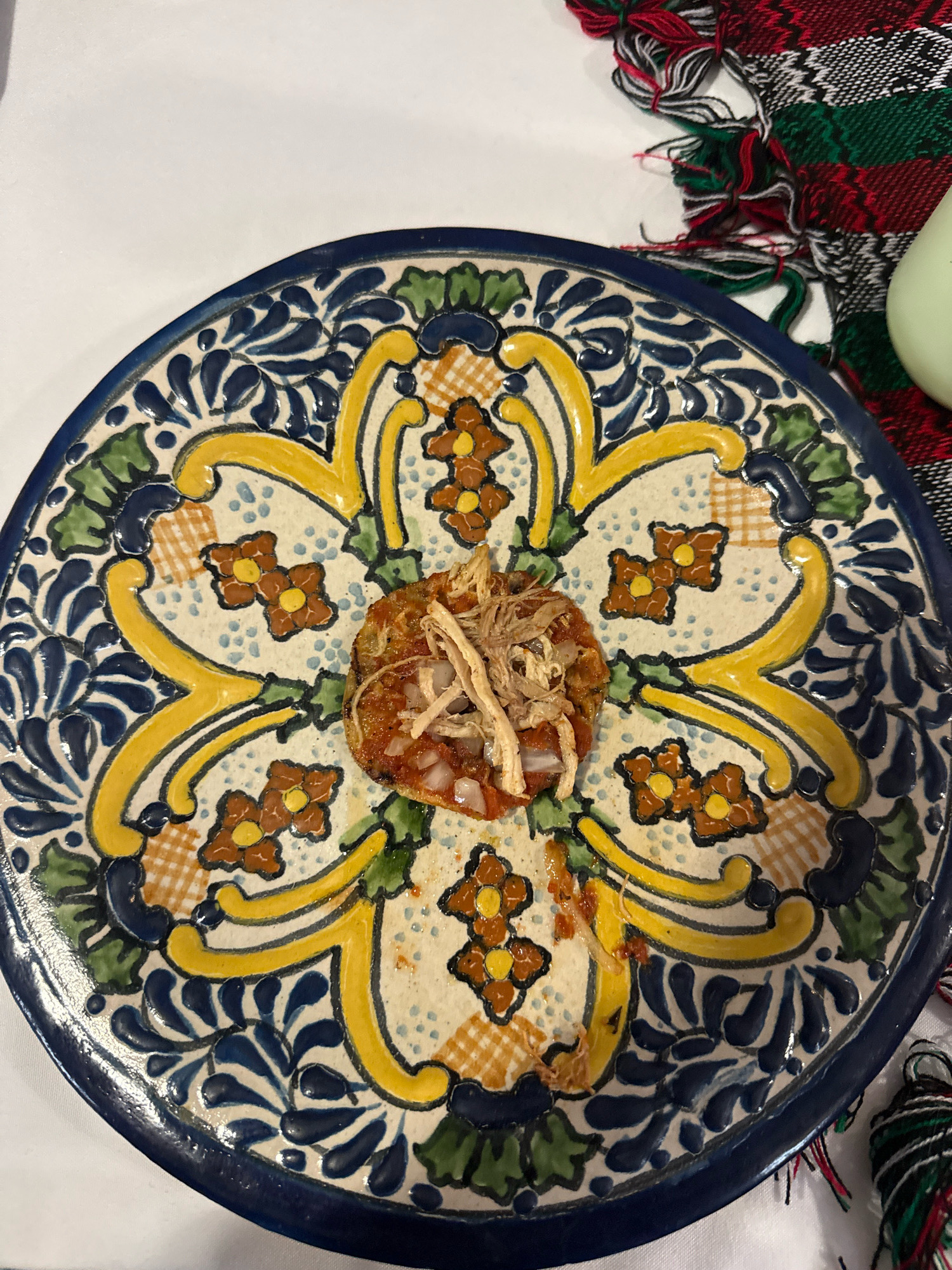 A close-up, overhead shot shows a single serving of food atop a decorative Talavera plate. The plate is white with a cobalt blue rim and features a colorful, floral pattern in yellow, green, and brown. The food, a small mound of shredded meat and finely diced onions in a red sauce, is positioned in the center of the plate, with a sprinkle of what appears to be thinly sliced dried chiles or herbs on top. The plate sits on a white surface, partially obscured by a festive, plaid-patterned cloth with red, green, and black threads and fringe. This scene is likely a photograph taken in a restaurant or home in the city of Puebla, Mexico, known for its distinctive Talavera pottery and traditional cuisine.