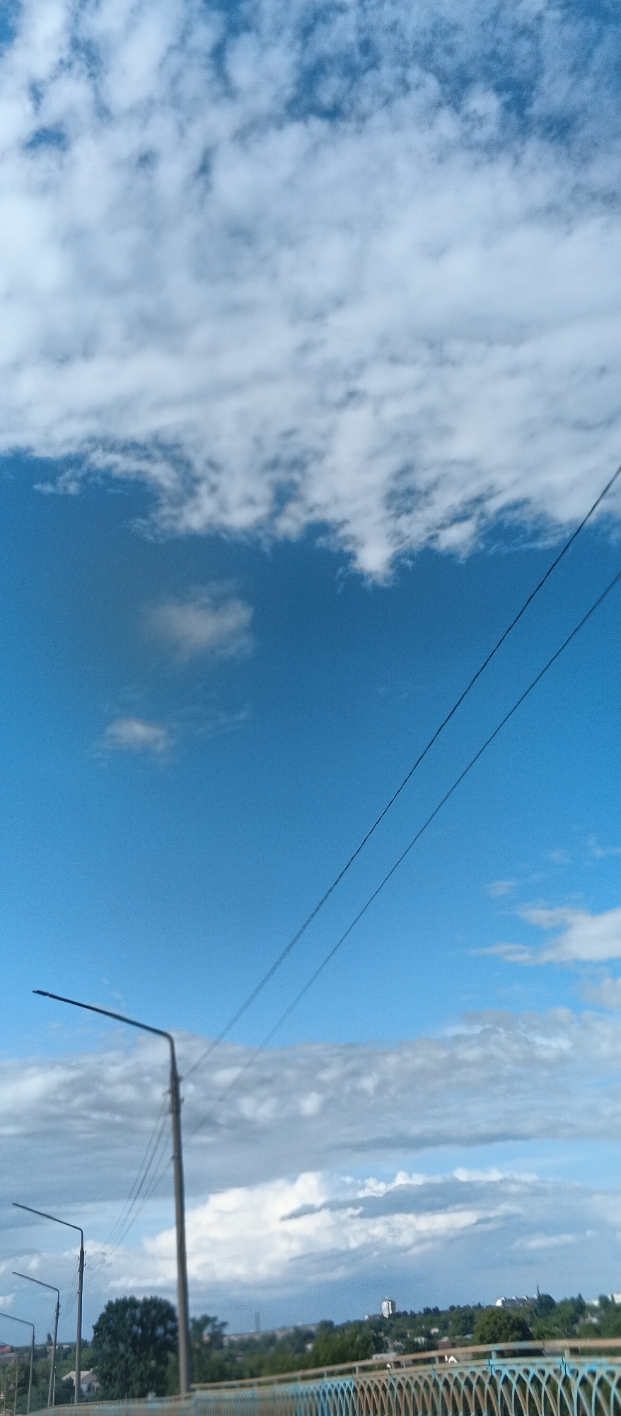 A vertical, medium shot captures a bright blue sky with scattered white clouds. Two dark power lines stretch diagonally across the upper right quadrant of the frame. In the lower half, a street lamp stands to the left of center, its pole and arm clearly visible against the sky. A second, shorter street lamp is positioned to the far left. Below these, a blurred landscape of green trees and buildings is visible, suggesting an urban or suburban environment. A light blue and orange railing runs along the bottom of the image, hinting at the perspective being from a bridge or elevated walkway. The overall scene is illuminated by daylight, with the sky being the dominant feature.