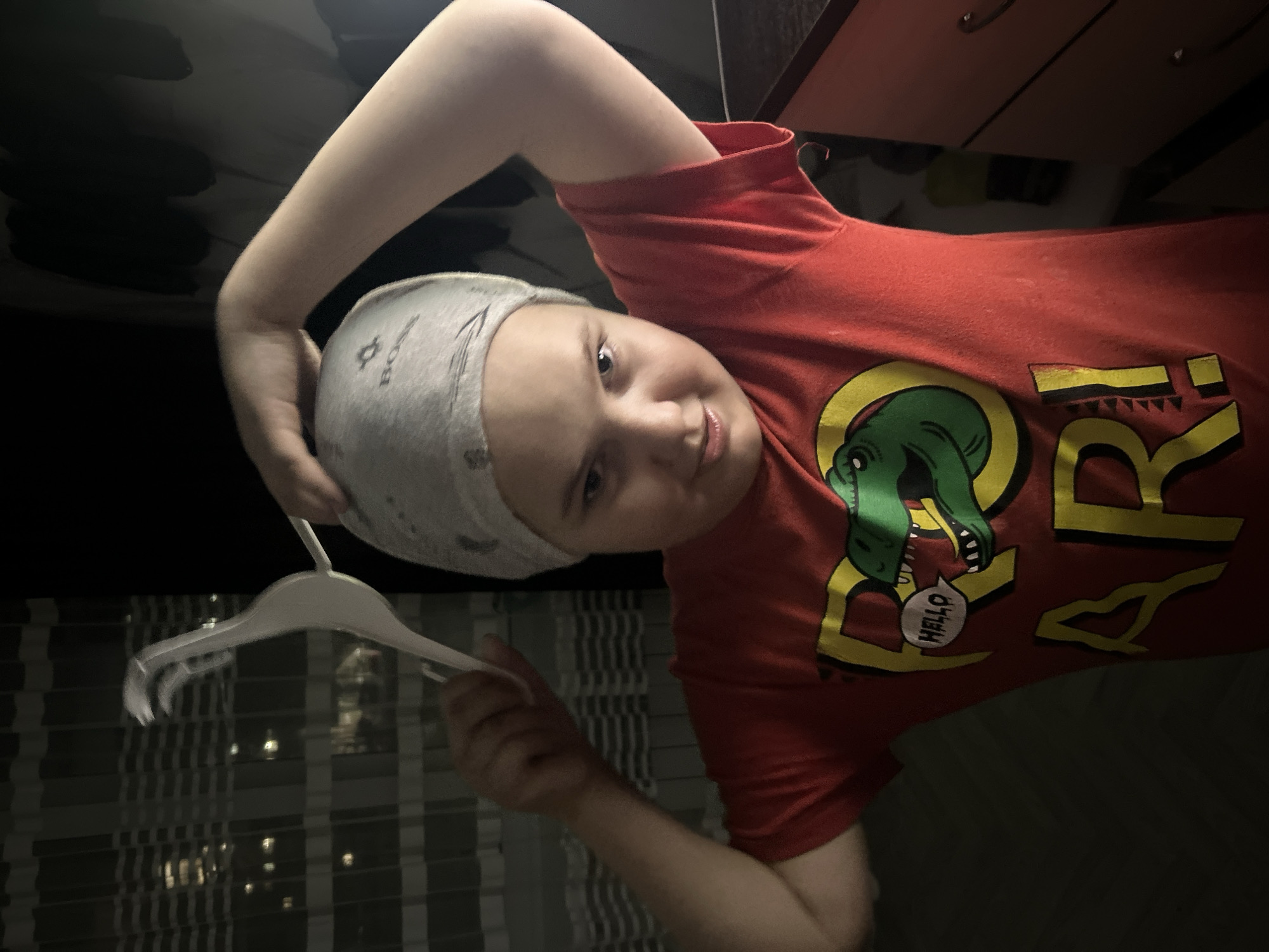 A child with a grey head covering is holding a white hanger in their left hand. They are wearing a red t-shirt with a dinosaur graphic. The background shows a darkened room with a view of a building with many windows. The location is implied to be Chaikovsky, Russia.