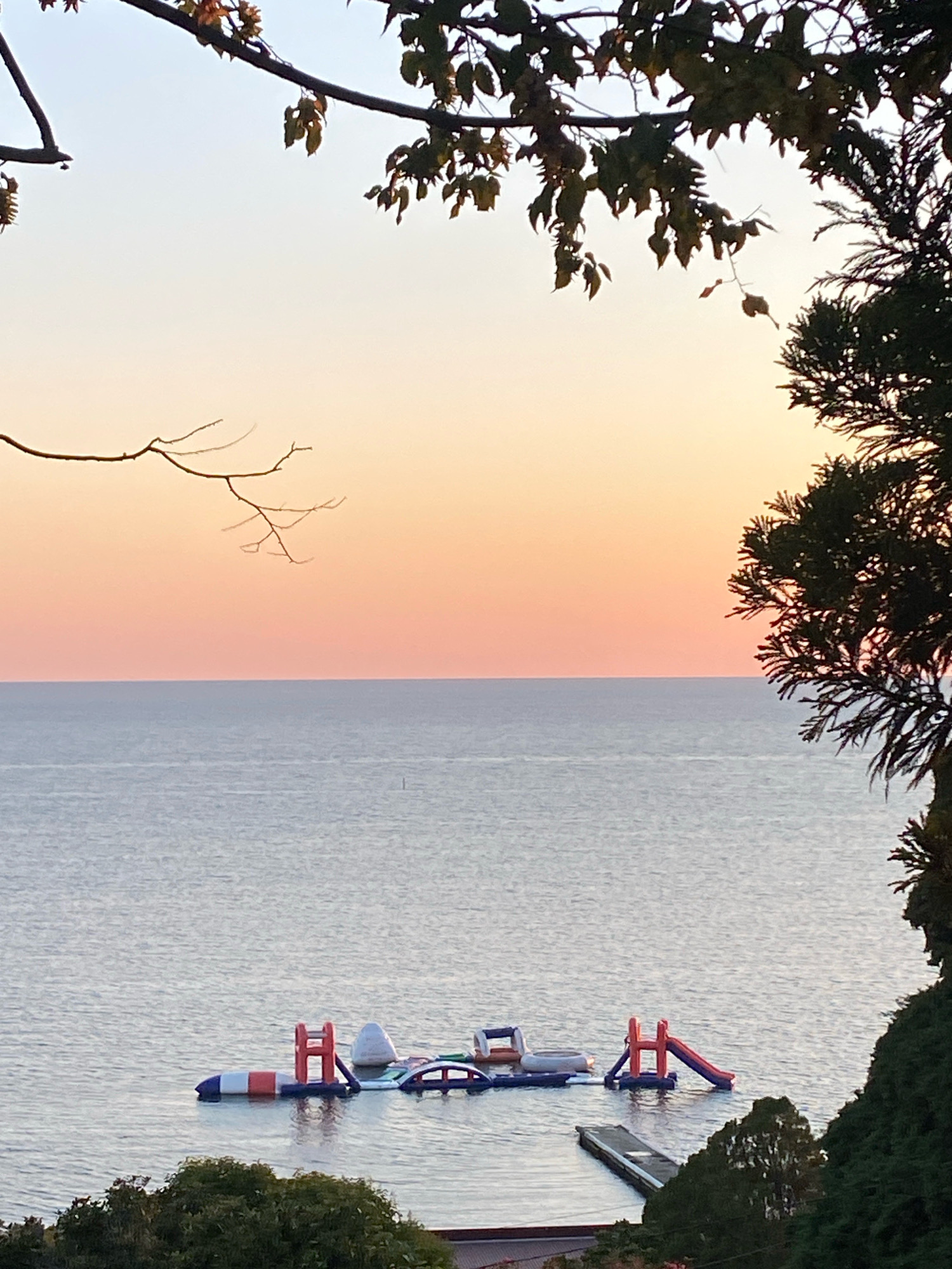 This image shows a tranquil scene of a body of water, likely the Black Sea, near Sochi, Russia, at sunset. The sky exhibits a gradient of soft oranges and pinks, transitioning to a pale blue towards the top. The water is calm and appears to reflect the colors of the sky. In the middle of the water, there is an inflatable water park, with various colorful structures including slides, a climbing wall, and platforms. The park is partially submerged, suggesting it's an attraction for recreational activities. In the foreground, lush green trees and branches frame the view, with some trees also visible on the right side of the image, partly obscuring the water. A wooden dock extends from the shore into the water on the right side of the frame. The overall atmosphere is peaceful and serene, typical of a coastal landscape during the evening. There are no people visible in the image.