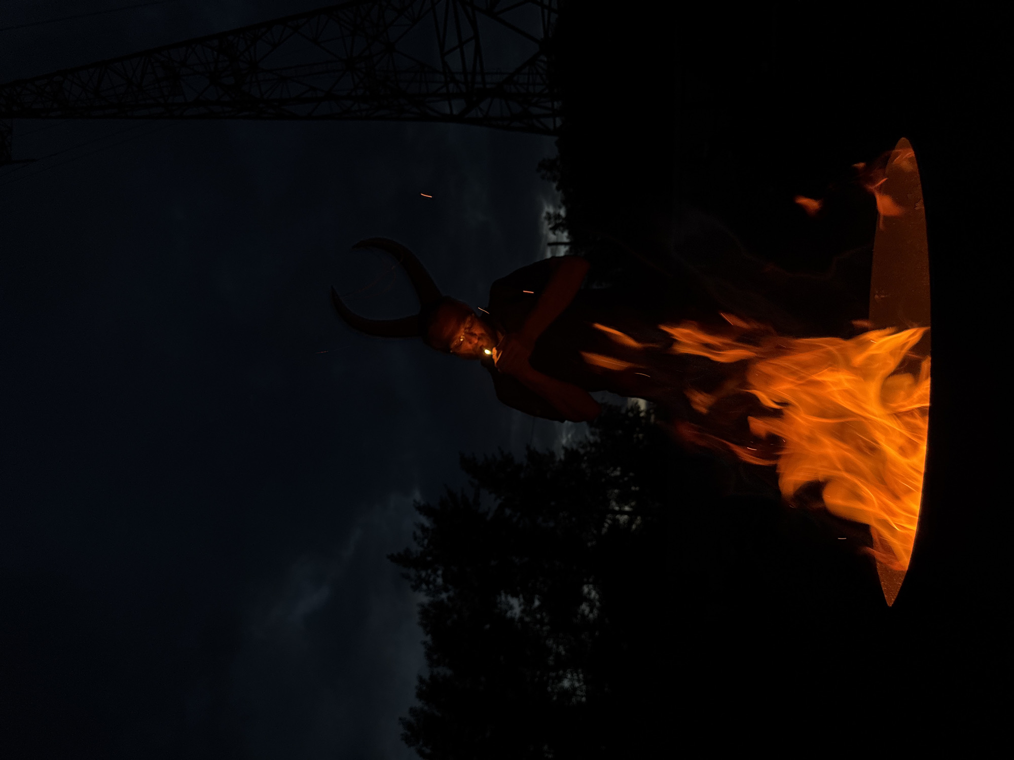 In Chaikovsky, Russia, on a dark night, a person stands by a fire pit, holding a lighter to ignite kindling. The person wears a costume with large, curved horns, resembling those of the Marvel character Loki. The fire casts an orange glow on their face and the surrounding flames. In the background, silhouetted trees and a tall, lattice-like tower, possibly a power line pylon, are visible against a cloudy, dark sky. The scene is lit primarily by the fire, creating a dramatic, low-light atmosphere.
