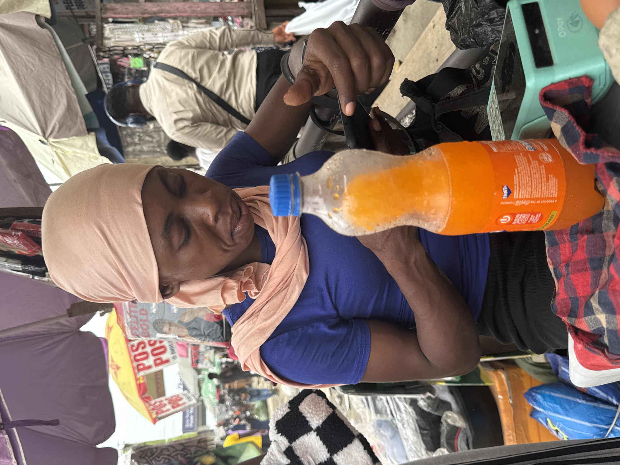 A Black woman wearing a pink headwrap and a blue t-shirt is seated behind a stall in an outdoor market, likely in Lekki, Nigeria. She is looking down at her mobile phone, which she is holding in her right hand. Her left arm is resting on the table in front of her. A bottle of Fanta orange soda with condensation on it stands to her right, next to a turquoise OPay point-of-sale device. A red and black checkered cloth is draped over the table. In the background, the market appears busy with other stalls and people, including a man wearing a light brown shirt and headphones. Umbrellas and awnings provide shade over the market.