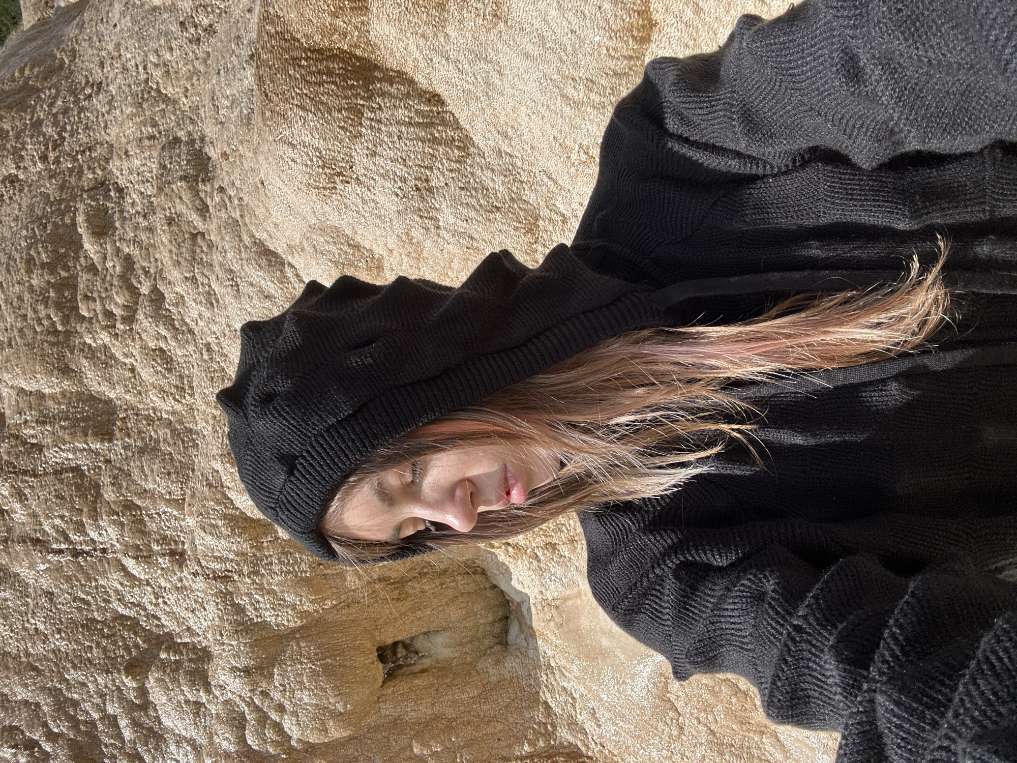 A young woman with light brown hair, some of which has pink highlights, is pictured from the chest up, wearing a black knit hooded sweater. She is looking down and to her left, with her eyes closed. The background is a textured, light brown rock formation. The location is not identifiable as Almasiani, Georgia.
