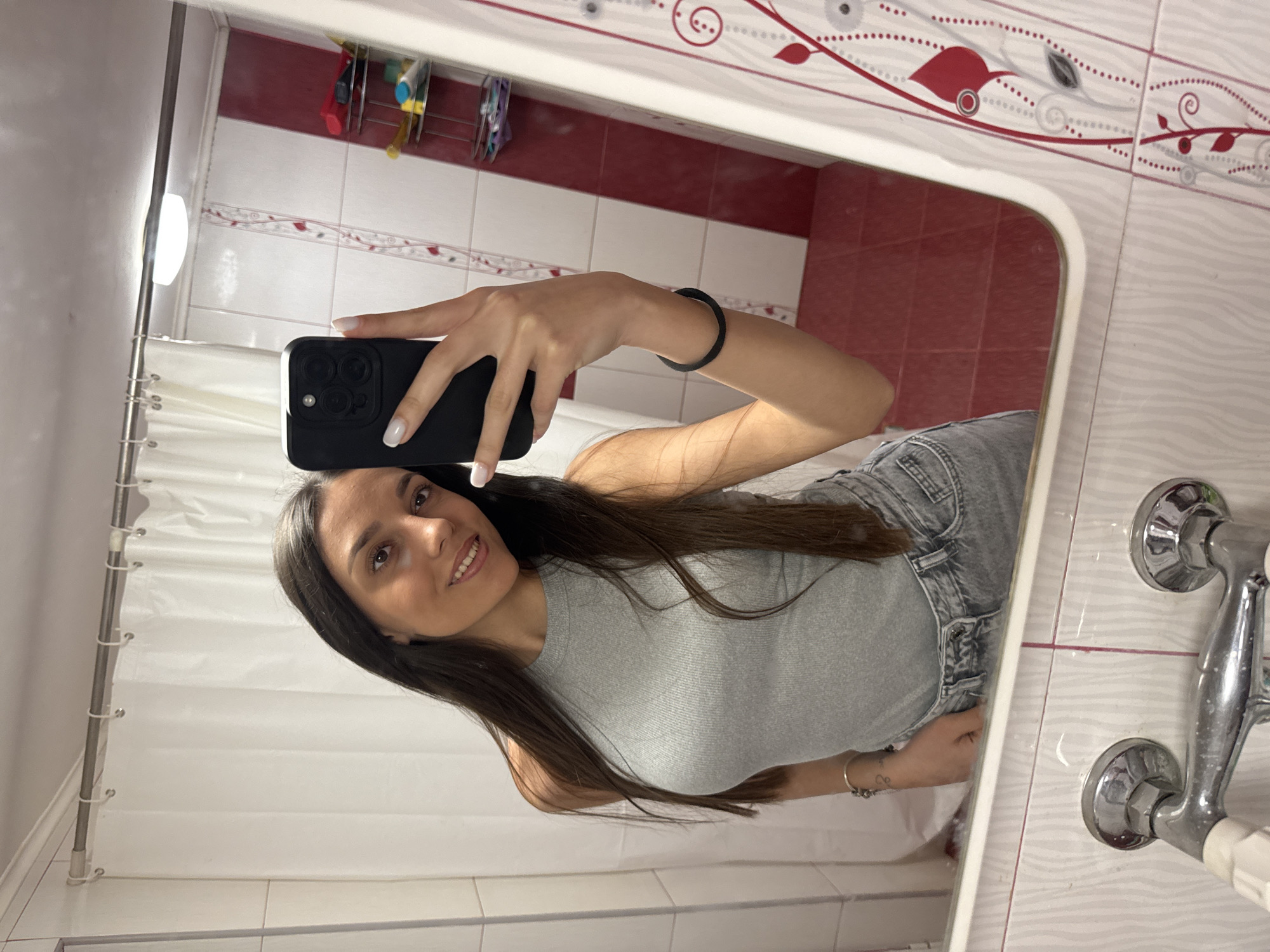 A young woman is taking a selfie in a bathroom, visible in a mirror. She's holding a black smartphone in her right hand. She's wearing a gray tank top and jeans. She has long, dark brown hair and is smiling slightly. The bathroom has a white shower curtain and white and red patterned tiles on the wall. A faucet is visible to the right side of the mirror. The specific location in Drenovets, Bulgaria, cannot be determined.
