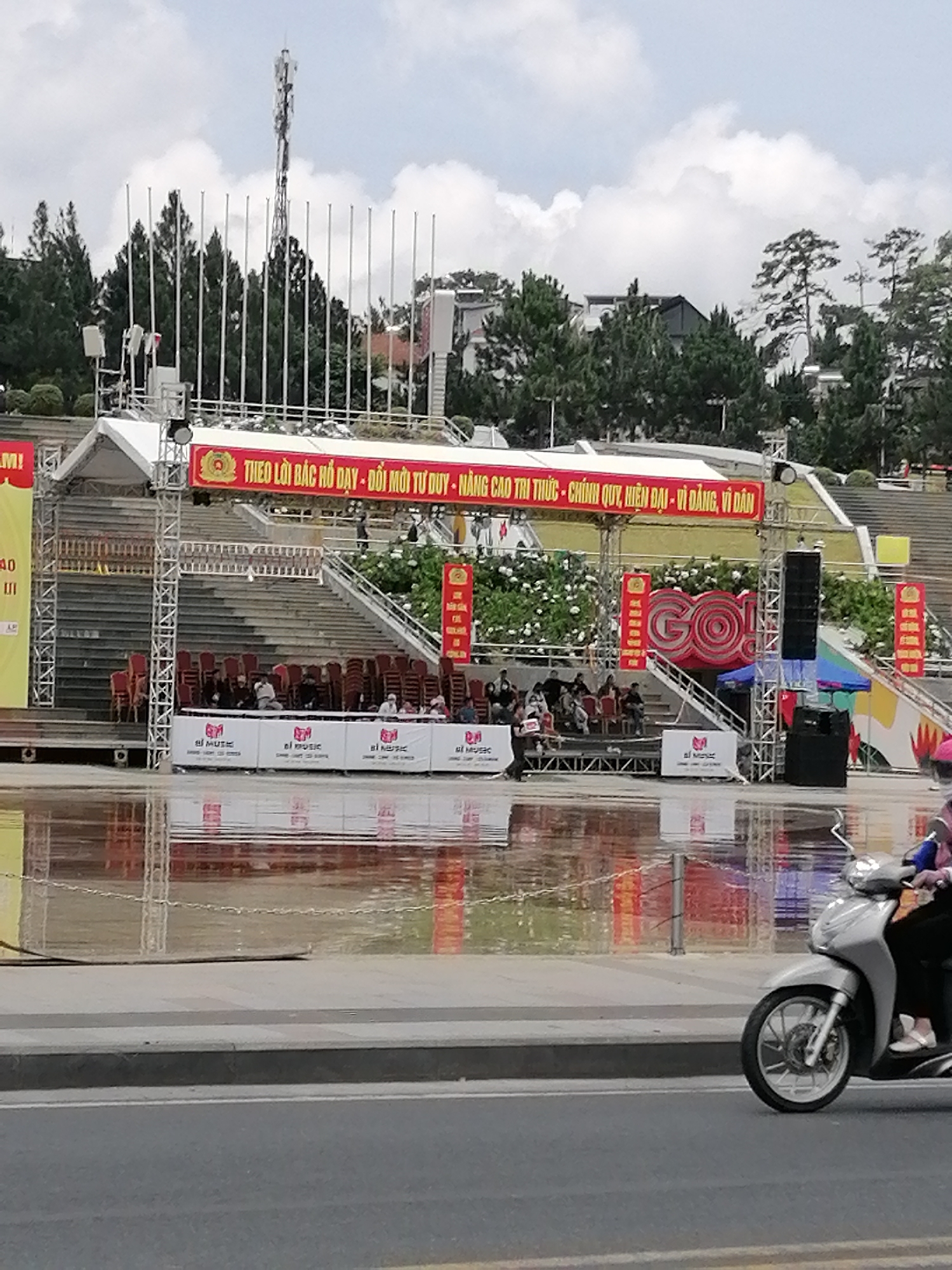 The image shows an outdoor event setup in Da Lat, Vietnam. In the background, there are tiered stadium seats with many empty chairs. A large red banner with Vietnamese text hangs across the top of the stage area. The banner includes a logo with what appears to be the Vietnamese seal. Below the banner, a stage structure is visible, with scaffolding and speakers. There are also vertical red banners with Chinese characters on either side of a large, decorative "GO!" sign. In front of the stage, there is a reflective surface, possibly water or a wet pavement, which mirrors the structures above. Several white banners with logos are also placed along the edge of this reflective area. In the foreground, a person is riding a silver scooter on a paved road. The overall setting suggests a public gathering or performance space.
