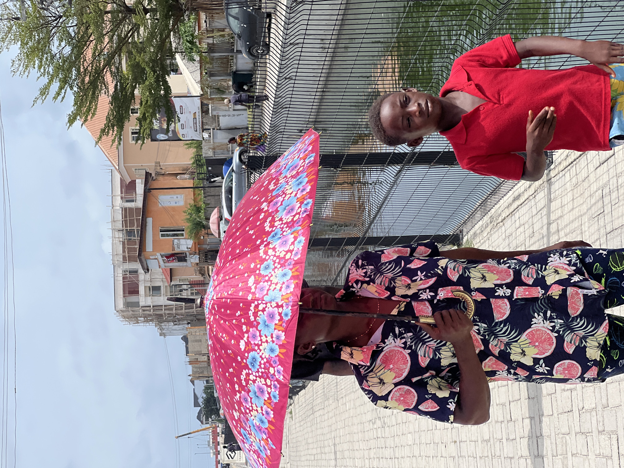 A medium shot captures a person holding a red floral umbrella alongside a child in Igboefon, Nigeria. The person is wearing a short-sleeved button-up shirt patterned with citrus fruit and tropical leaves and also wears glasses. They are holding the umbrella, which has a dark handle, to provide shade. The child, wearing a red polo shirt, is positioned next to a black metal fence that overlooks a water source. In the background, there are multi-story buildings, some under construction with scaffolding, and parked vehicles. The sky is overcast. The ground appears to be paved.