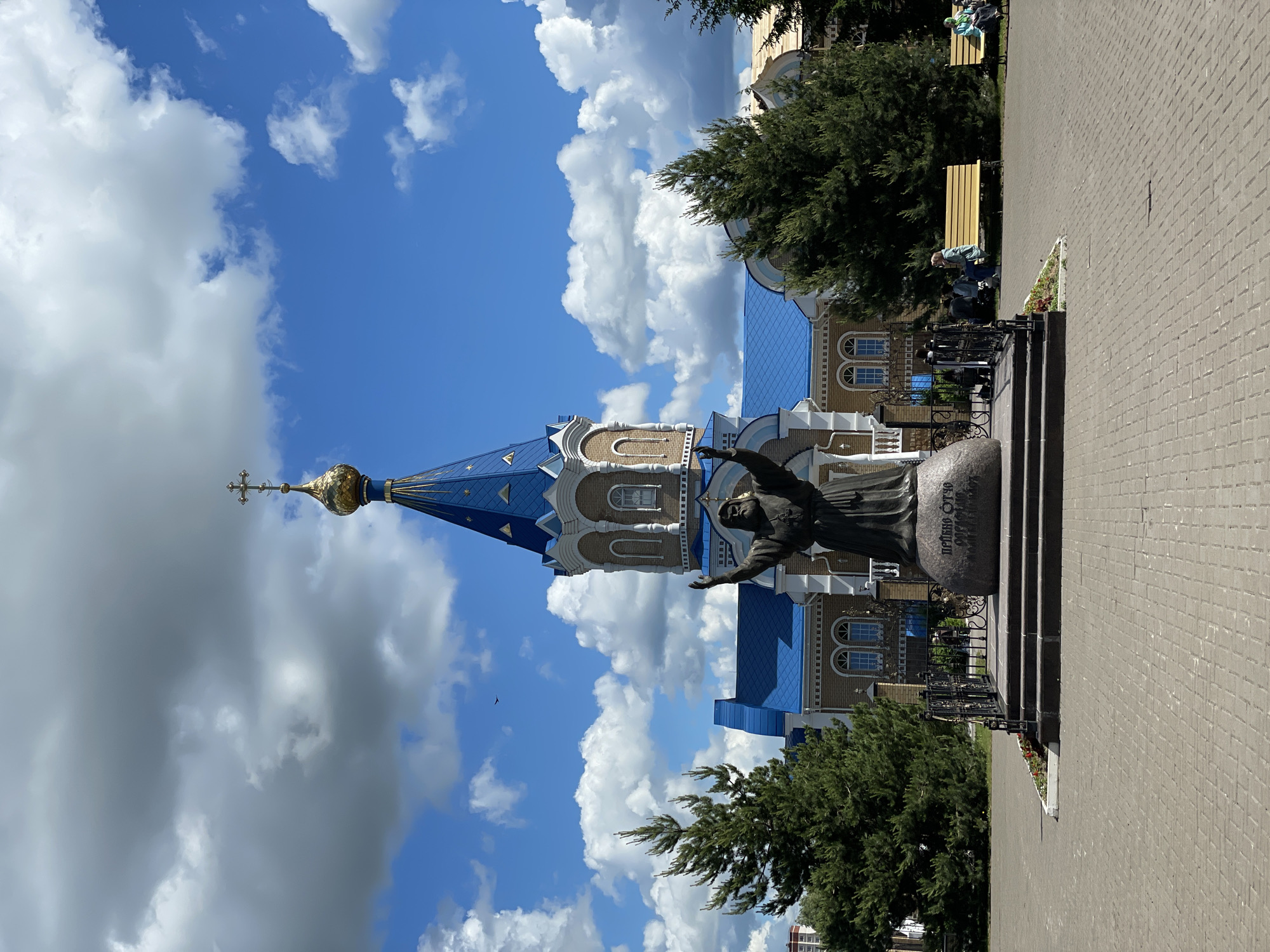The image depicts a sunny day scene in Izhevsk, Russia, featuring a church with a distinctive blue roof and a golden dome. A statue of a figure with outstretched arms stands in front of the church. A paved walkway extends to the right of the statue. Benches are visible along the sidewalk. The sky is predominantly blue with scattered cumulus clouds. The scene is well-lit, and the image is vertically oriented.