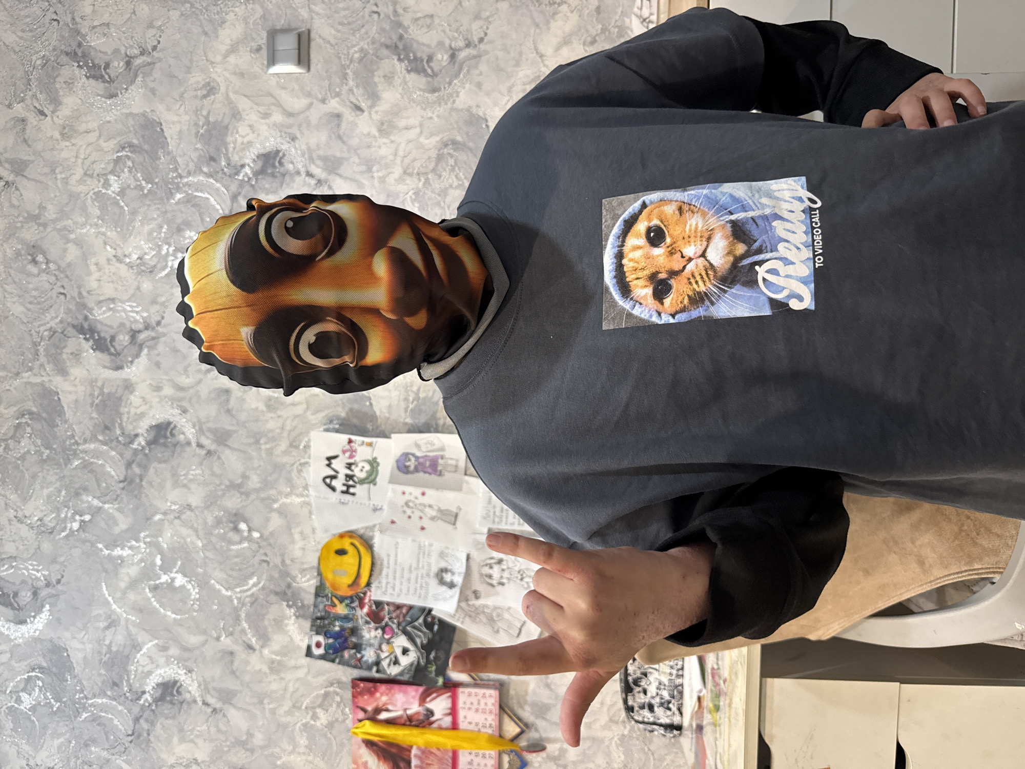 A person wearing a mask and a dark-colored sweatshirt with a cat graphic is standing against a wall with a mottled grey and white pattern. The person is making a hand gesture. On the wall below are various drawings and stickers. The location is in a room somewhere in Rusanovo, Russia.