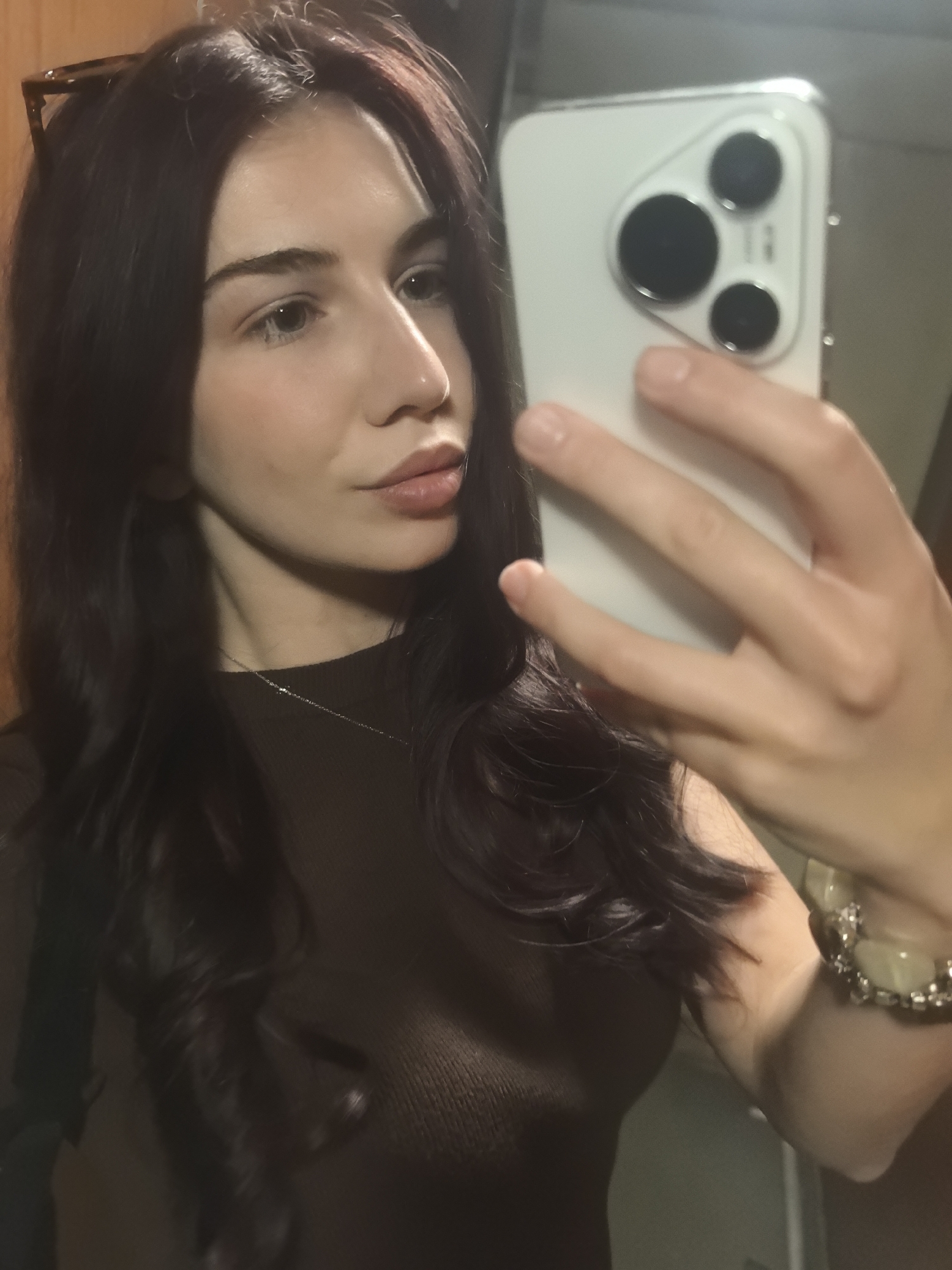 A woman is taking a selfie with a modern smartphone. The phone obscures part of her face. She has long, dark hair with a slight wave. She's wearing a dark, possibly brown top. A necklace is visible around her neck. Sunglasses are perched on her head. A bracelet adorns her wrist. The background is blurry, suggesting an indoor setting. The location in the city is Unknown, Unknown.