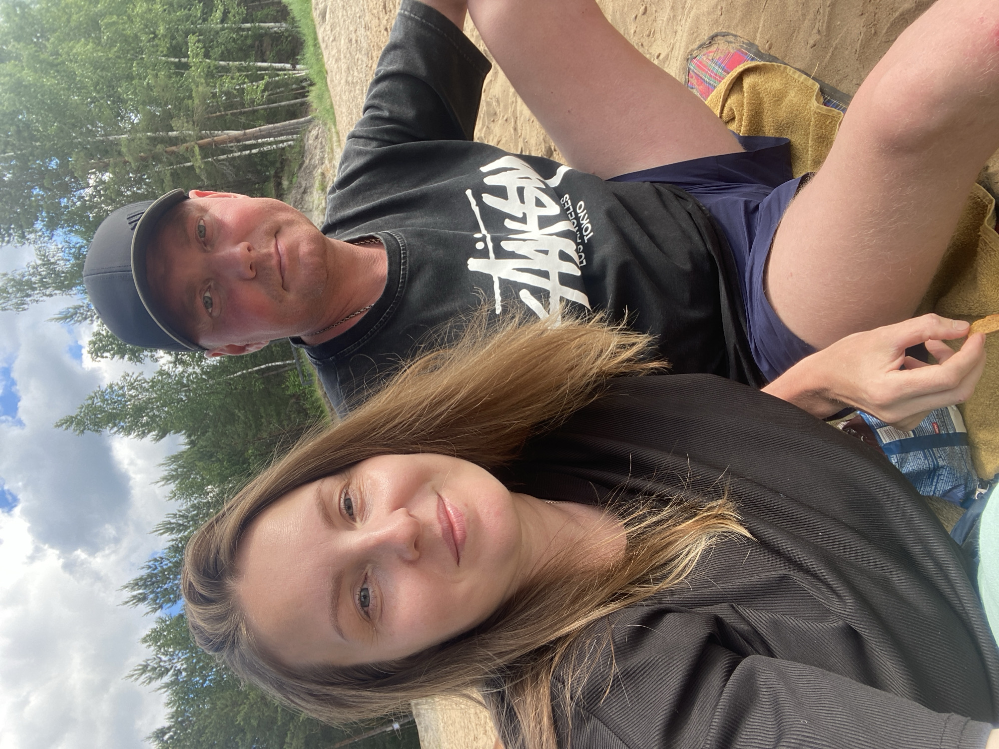 The image shows a couple sitting on a sandy beach with trees in the background. The woman on the left is wearing a black ribbed shirt and looking at the camera with a slight smile. The man on the right is wearing a black t-shirt with a white logo and a black baseball cap, also looking towards the camera. He has a chain necklace visible. They are both sitting close together, with the man's arm resting on his leg and his hand near a bag of chips. The environment suggests a warm, sunny day, likely a recreational area. There are clouds in the sky and the trees appear to be pine and birch trees.