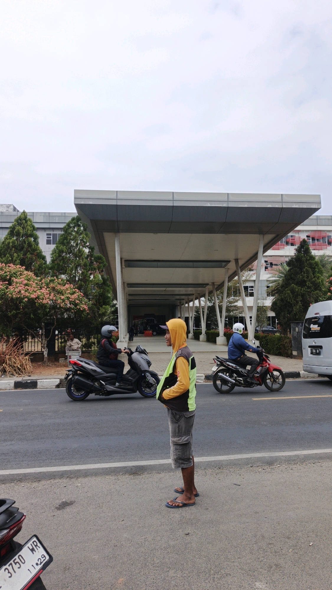 In Pangandaran, Indonesia, a man wearing a yellow hooded jacket with a lime green vest over it and grey cargo shorts stands on a paved road, facing right. He has on dark flip-flops. Behind him, a dark grey scooter passes by with a person wearing a helmet and a dark jacket riding it. Further in the background, a red and black motorcycle is ridden by a person in a blue shirt and helmet. To the far right, a white van is parked on the side of the road. The building in the background is modern with a grey, cantilevered roof supported by white columns. Trees and foliage are visible to the left of the building. The sky is overcast.