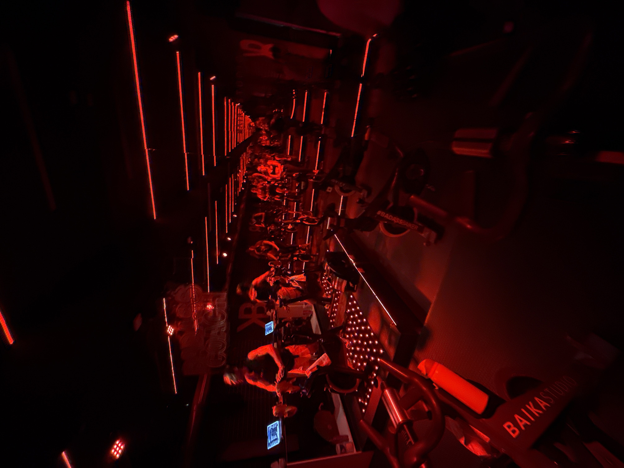 The image depicts the interior of a fitness studio, presumably in Puebla, Mexico, based on the studio name "BAIKA STUDIO" visible in the foreground. The scene is illuminated by a predominantly red lighting scheme, which casts a dark and intense atmosphere. Rows of stationary bikes, occupied by indistinct figures pedaling, stretch into the distance. Vertical red light fixtures line the walls, contributing to the tunnel-like appearance of the space. The focus is on the rhythmic action of the cyclists engaged in a group fitness session.