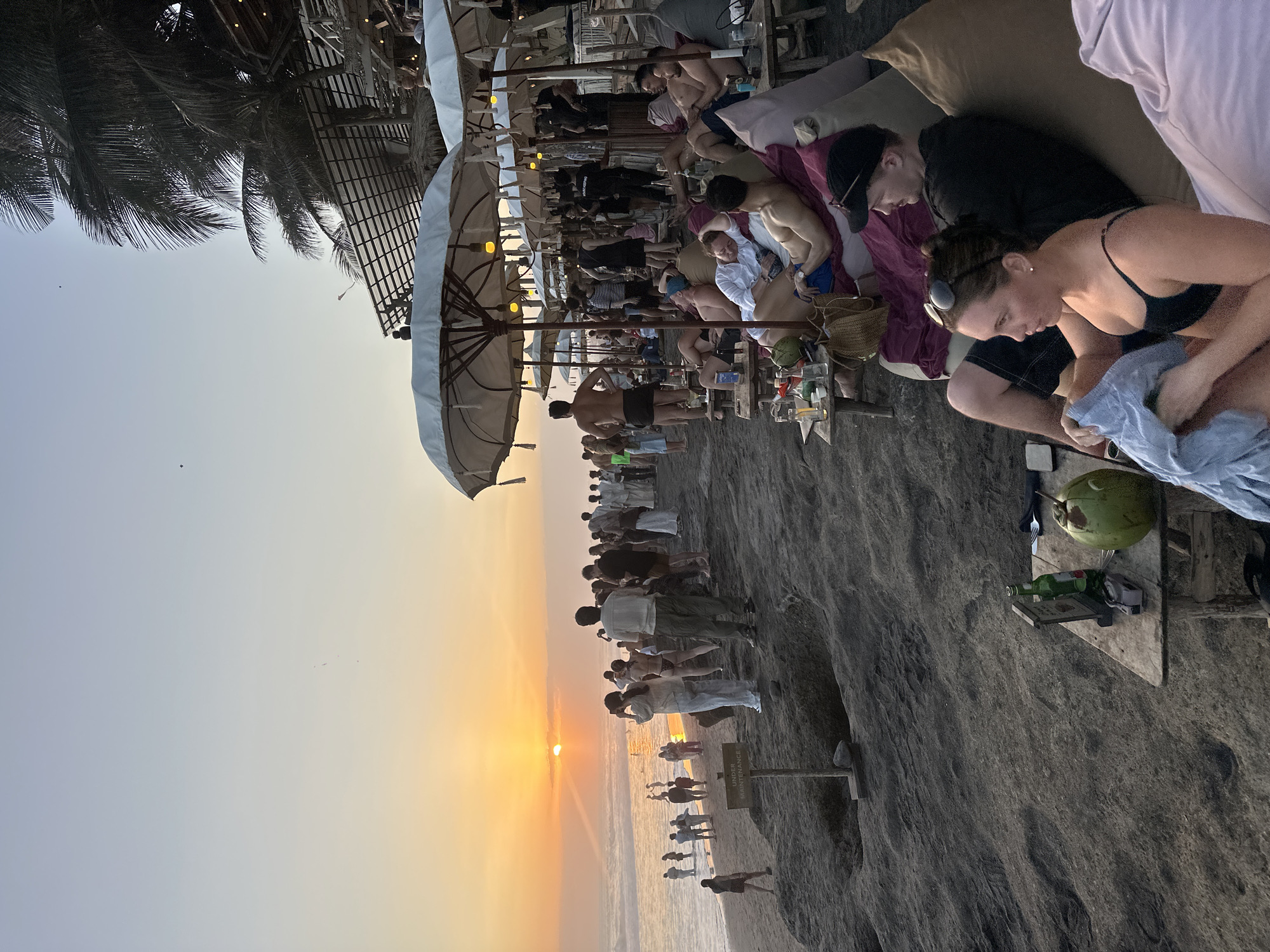 A crowded beach at sunset features people relaxing on loungers and standing near the water. In the foreground, a woman in a black bikini is seated on a large cushion, holding a light blue cloth. Next to her, a man wearing a black t-shirt and a baseball cap is also seated. A wooden table in front of them has a green coconut, a bottle of beer, and a small sign on it. Further back, many people are standing on the dark sand beach, some watching the sunset over the ocean. Several white umbrellas and thatched-roof structures suggest a beachfront bar or restaurant. Palm trees are visible in the upper right portion of the image. The sun is low on the horizon, casting a warm glow over the scene.