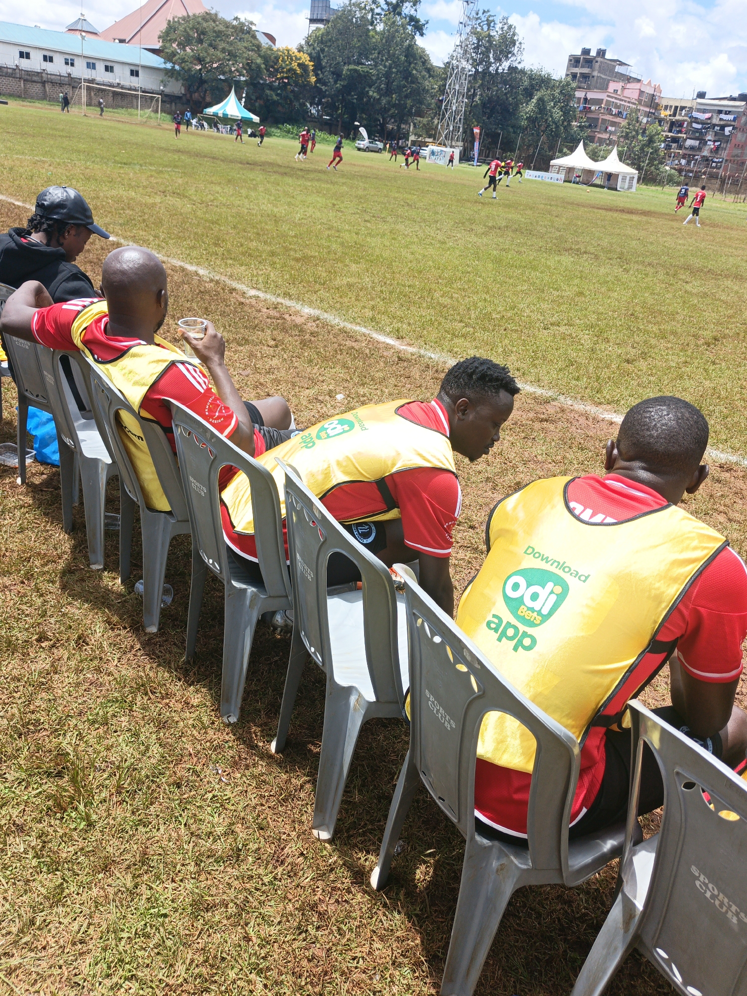 The image depicts a soccer field in Nairobi, Kenya. A match is in progress, with players visible on the green field under a partly cloudy sky. In the foreground, four individuals sit in gray plastic chairs along the sidelines. All are wearing red and yellow vests, and they are presumably associated with one of the soccer teams. The vests feature a "Download Odi Bets App" logo. The backdrop includes buildings typical of Nairobi's urban landscape. A water tower is visible in the background as well.