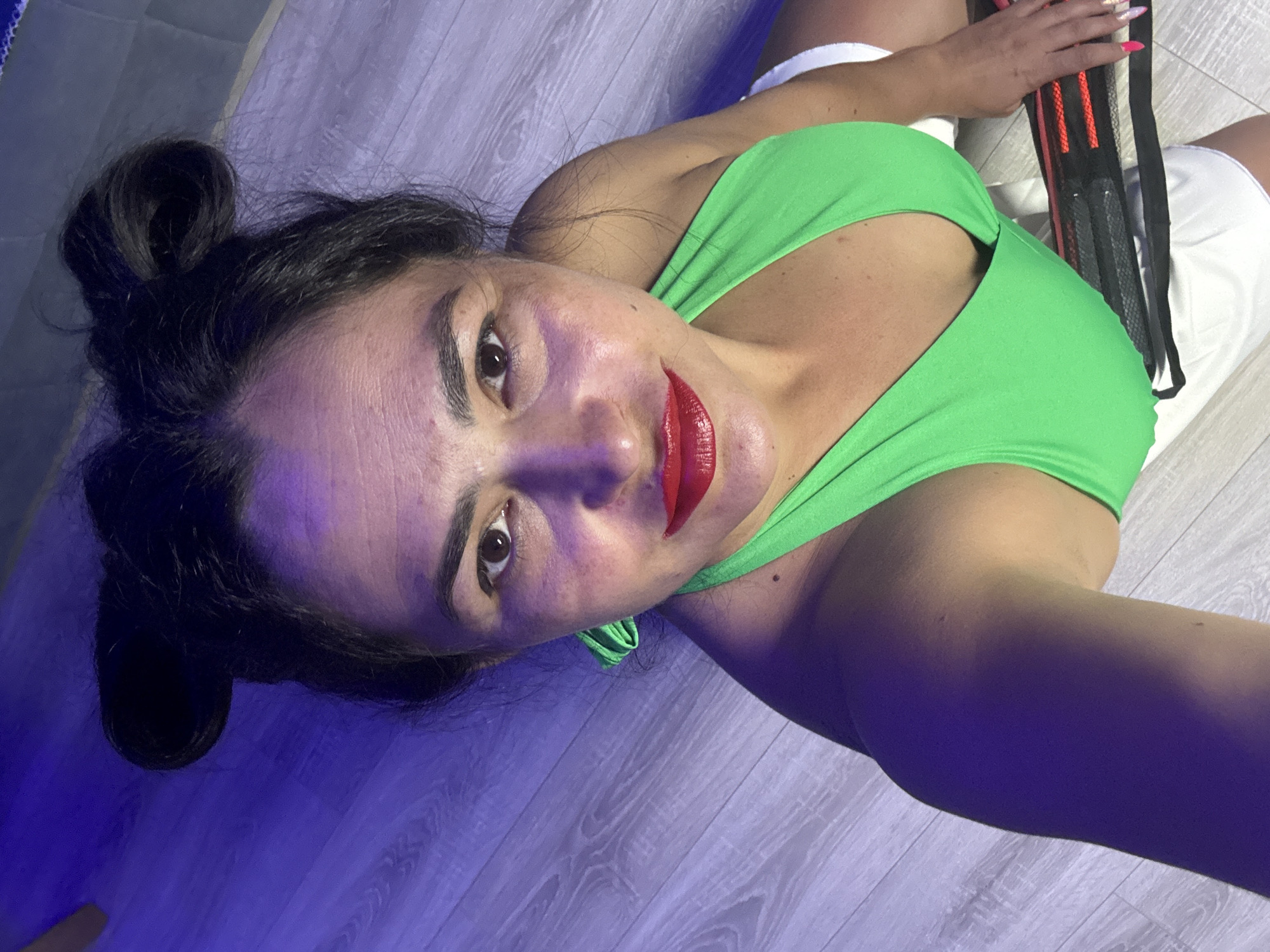 A woman is captured in a selfie-style shot, lying or sitting on a light-colored wooden floor in an indoor setting, likely in Kauchuk Stantsiya, Russia. She looks directly up at the camera with a neutral to slightly smiling expression. She has dark hair tied back in a messy bun, dark eyebrows, and is wearing bright red lipstick.
She is dressed in a vibrant green athletic top or sports bra and white shorts or athletic bottoms. Her right hand is visible, holding a black strap with orange mesh details, which appears to be part of a resistance band set, suggesting she is either working out or taking a break from a fitness activity. Her fingernails are manicured with pink and white polish.
The scene is illuminated by strong artificial blue and purple lighting, which casts distinct hues and shadows on her skin and the wooden floor. The floor has a visible grain pattern. The overall atmosphere suggests a gym, studio, or personal workout space. There is no visible text in the image.
