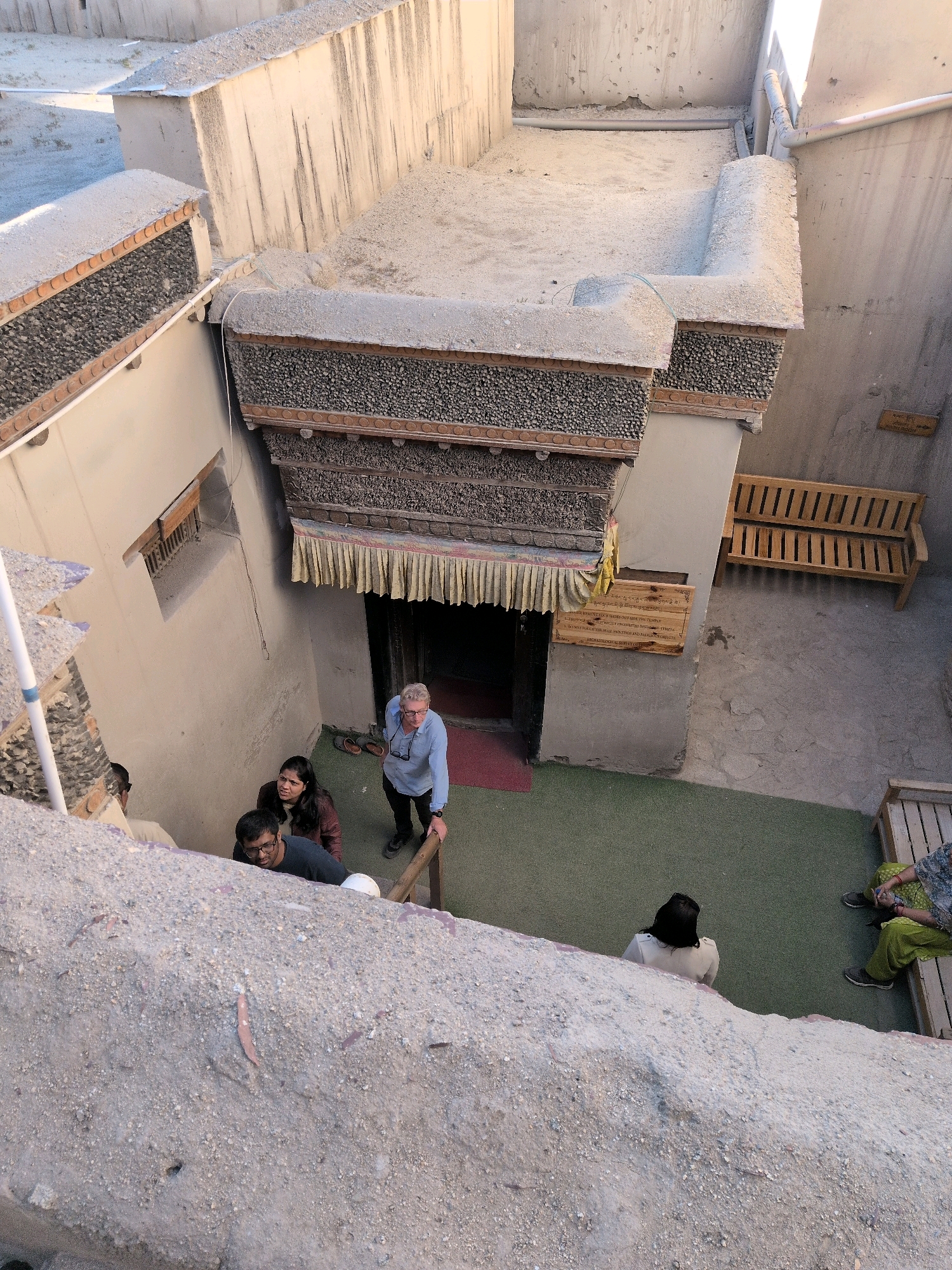The image shows an outdoor courtyard of an old building in Leh, India. A group of people are seen standing on a set of stairs, some looking up. In the background, there is a wooden bench and some signage on the wall. The architecture appears traditional, with textured walls and decorative elements. The overall scene suggests a historical site or cultural landmark.