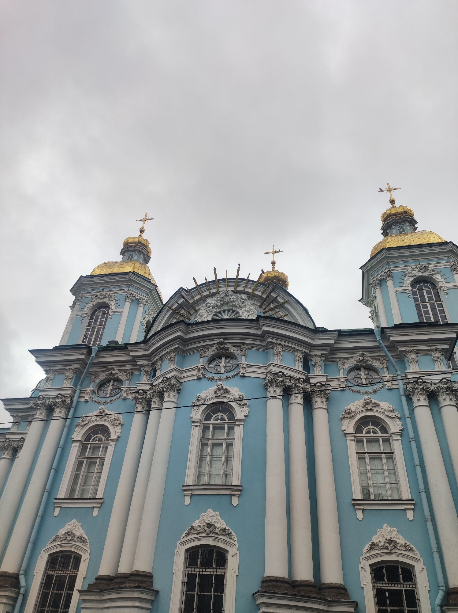 The image shows a low-angle shot of a blue and white Baroque church with golden domes and crosses against a cloudy sky. The church features tall, white columns and ornate windows with decorative trim. The architectural style is characteristic of Saint Petersburg, Russia.