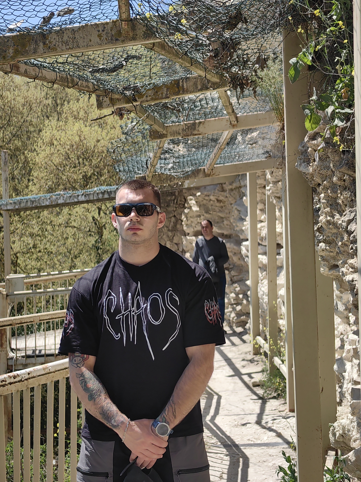 The image depicts a man wearing sunglasses and a black T-shirt with the word "CHAOS" printed on it. He has tattoos on his arms and is wearing a watch. He is standing on a pathway constructed with weathered white-painted metal railings and support posts, which may suggest an elevated walkway or bridge. Another man is walking in the background. The pathway is surrounded by a stone wall on the right and trees on the left, suggesting a park or garden setting. Above the man, there is a partially covered overhead structure made of metal beams and netting, which creates a shaded area. The overall environment appears to be outdoors on a sunny day. Based on the stone walls and the type of structure, the location might be within a historical park or garden in Varna, Bulgaria.
