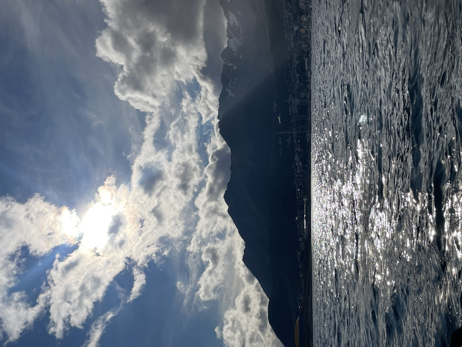 A striking image portrays a scene of nature's grandeur. The sky is partly cloudy with the sun partially obscured. Majestic mountains are visible behind the clouds. Water ripples in the foreground, reflecting sunlight and giving the surface a shimmering appearance. A coastline with buildings runs along the base of the mountains. The image is rotated 90 degrees to the left.