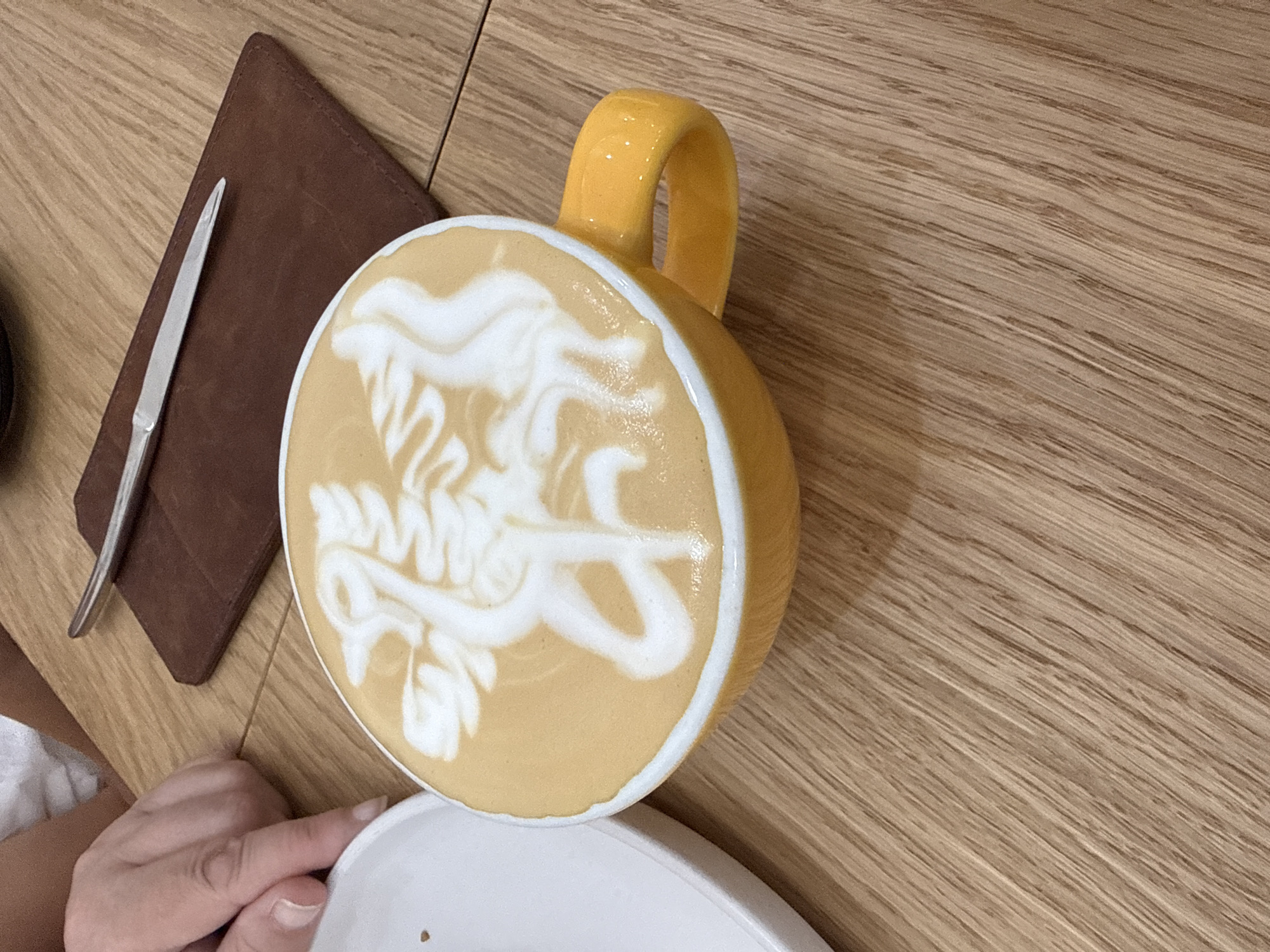 An overhead, medium shot captures a yellow ceramic mug filled with latte art on a light-brown wooden table. The latte art depicts a white mythical creature with wings, a beak, and a long, curved tail, etched onto the frothy surface of the coffee. To the left of the mug, a white plate is partially visible. In the upper left corner, a person's hand, with manicured fingernails, rests on the table, with fingers extended toward the white plate. A dark brown leather coaster lies to the right of the mug, with a silver utensil resting on top. The wooden table's grain runs diagonally across the frame.