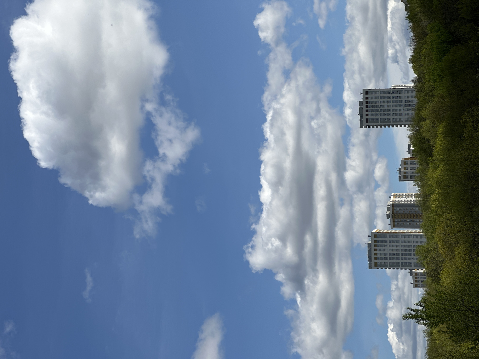 The image shows a bright blue sky with puffy white clouds. On the right side of the image, behind a line of green trees, stand several tall buildings of white or light-colored construction. It may be in the city of Nizhny Novgorod, Russia.
