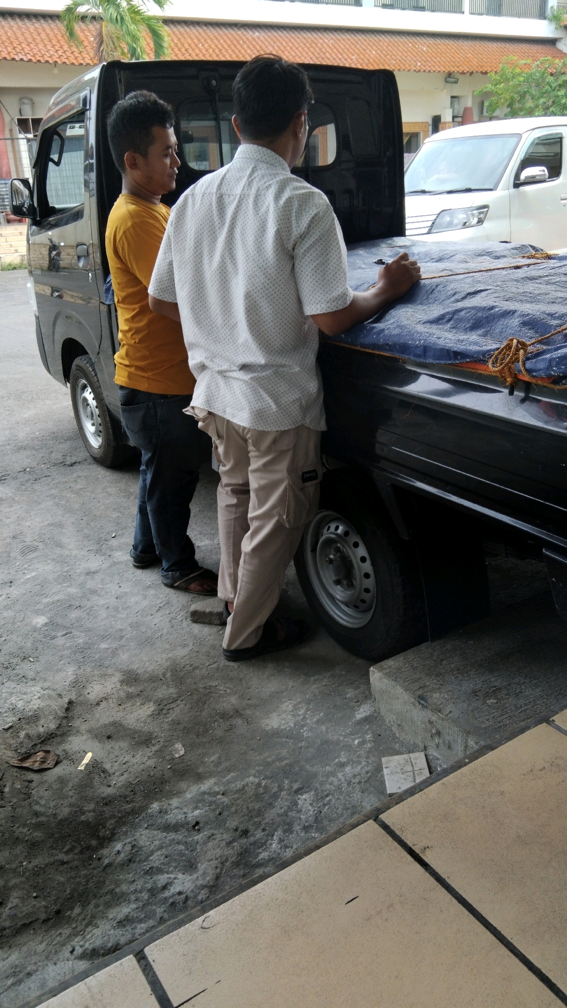 Two men are standing next to a black pickup truck covered with a blue tarp. The man on the left wears a yellow t-shirt and dark blue jeans. The man on the right wears a white collared shirt with a dotted pattern and light brown pants. The truck has a black bed with a blue tarp secured by rope. A white van is parked behind the truck, and a building with a tiled roof is visible in the background. The ground is a rough, concrete surface. The scene appears to be in an outdoor setting, possibly a service area or parking lot. The presence of a white van and the architectural style of the building suggest this could be in Pekalongan, Indonesia, a city known for its textile industry and coastal location.