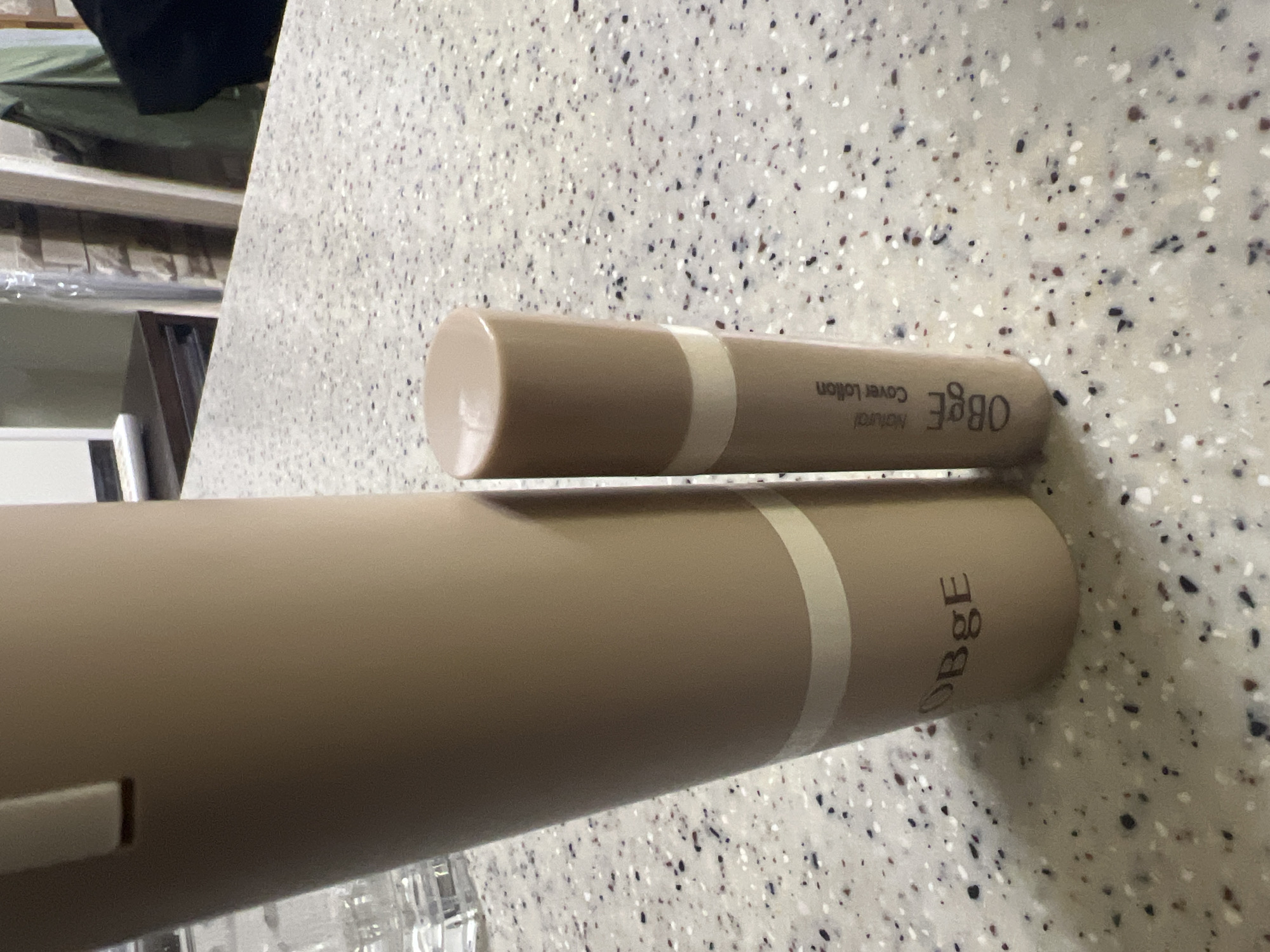 The image displays two cylindrical, beige-colored objects lying horizontally on a speckled light-colored countertop. One object is a slender tube, positioned above and slightly in front of a much larger, thicker tube. Both objects feature a lighter, cream-colored horizontal band near one end. The background is out of focus, revealing shelves or furniture.
The smaller tube has text printed along its side, which reads "Natural" and "Cover Lotion" in a darker font, followed by "OBgE" in a stylized capital font. The larger tube also has the brand name "OBgE" printed on its side. These appear to be cosmetic products.
The countertop surface is characterized by a light grey or white base with scattered black, grey, and reddish-brown specks, creating a textured appearance. No specific activity is taking place. The scene is lit from an unseen indoor source, creating subtle reflections and shadows. The time of day and weather are indeterminable. The image is rotated 90 degrees clockwise, presenting the horizontally oriented objects as if they are standing vertically in the frame. No emotions are conveyed.