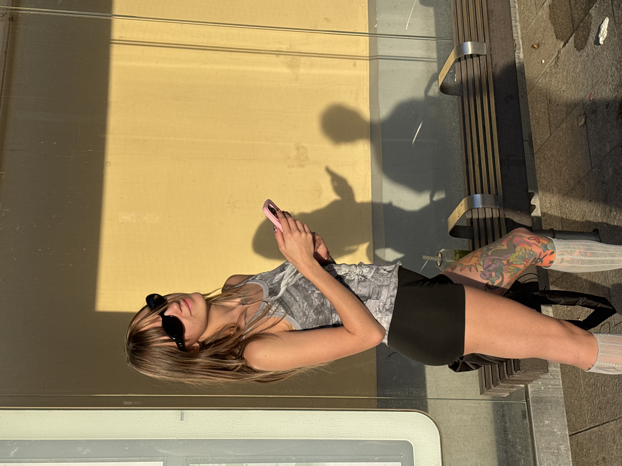 A young woman with long, light brown hair, wearing black sunglasses, a grey patterned tank top, black shorts, and white patterned socks, stands in a city setting and looks up and to her right. She holds a pink smartphone in her hands. Behind her, a yellow wall is illuminated by sunlight, casting shadows. A bench is visible in the background. The ground appears to be paved. There is a green drink in a plastic cup on the bench. The woman has extensive tattoos on her left leg. The scene is set in Moscow, Russia.