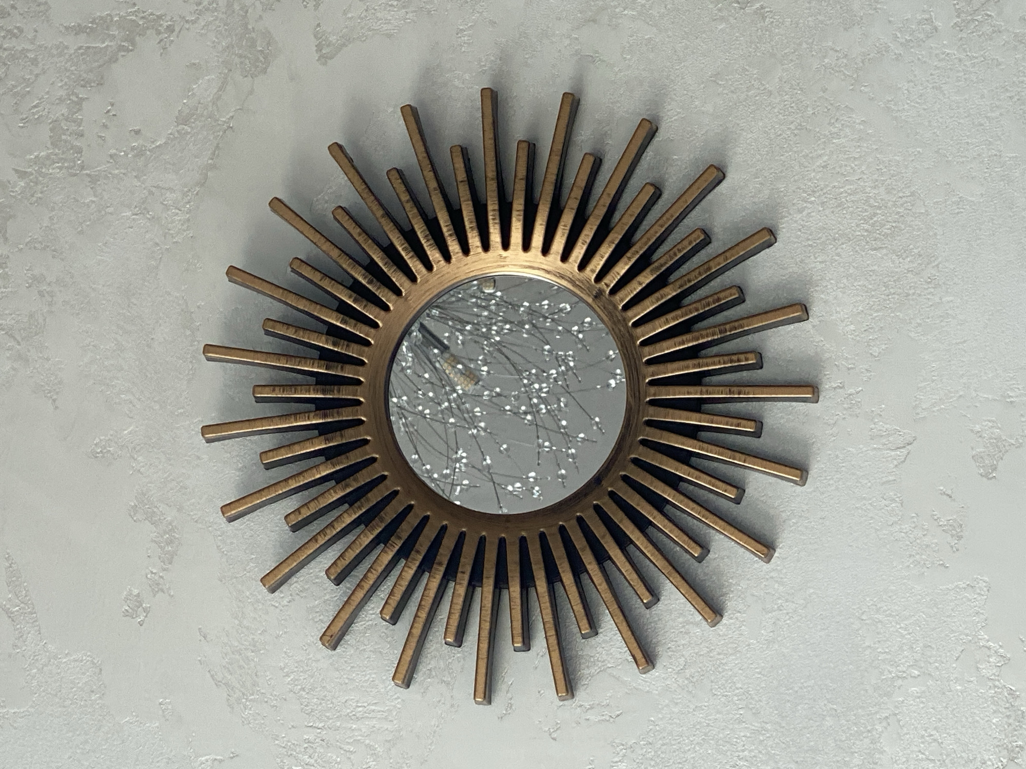 A sunburst mirror with a bronze frame is mounted on a textured white wall. The mirror reflects a light fixture with numerous small, white, bulb-like elements. The sunburst design consists of numerous thin, rectangular spokes radiating outwards from the central circular mirror. The wall has a rough, stucco-like finish. The scene is indoors and appears to be in a home or decorative space. There are no people or animals present.