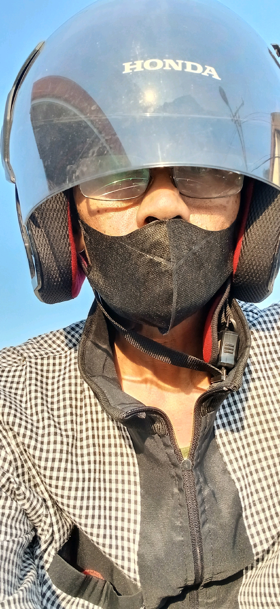A close-up, selfie-style, eye-level medium shot shows a person wearing a black and white checkered shirt and a black jacket. The person is also wearing a black face mask and a dark-colored helmet with "HONDA" visible on the front. The helmet has a clear visor, which reflects sunlight and a distant object. The person is wearing eyeglasses, which appear slightly cracked. The background is a clear blue sky. The scene appears to be outdoors, with no specific urban or natural landmarks to indicate the location within Samarinda, Indonesia. The lighting suggests it is daytime, possibly late afternoon due to the angle of the sun.