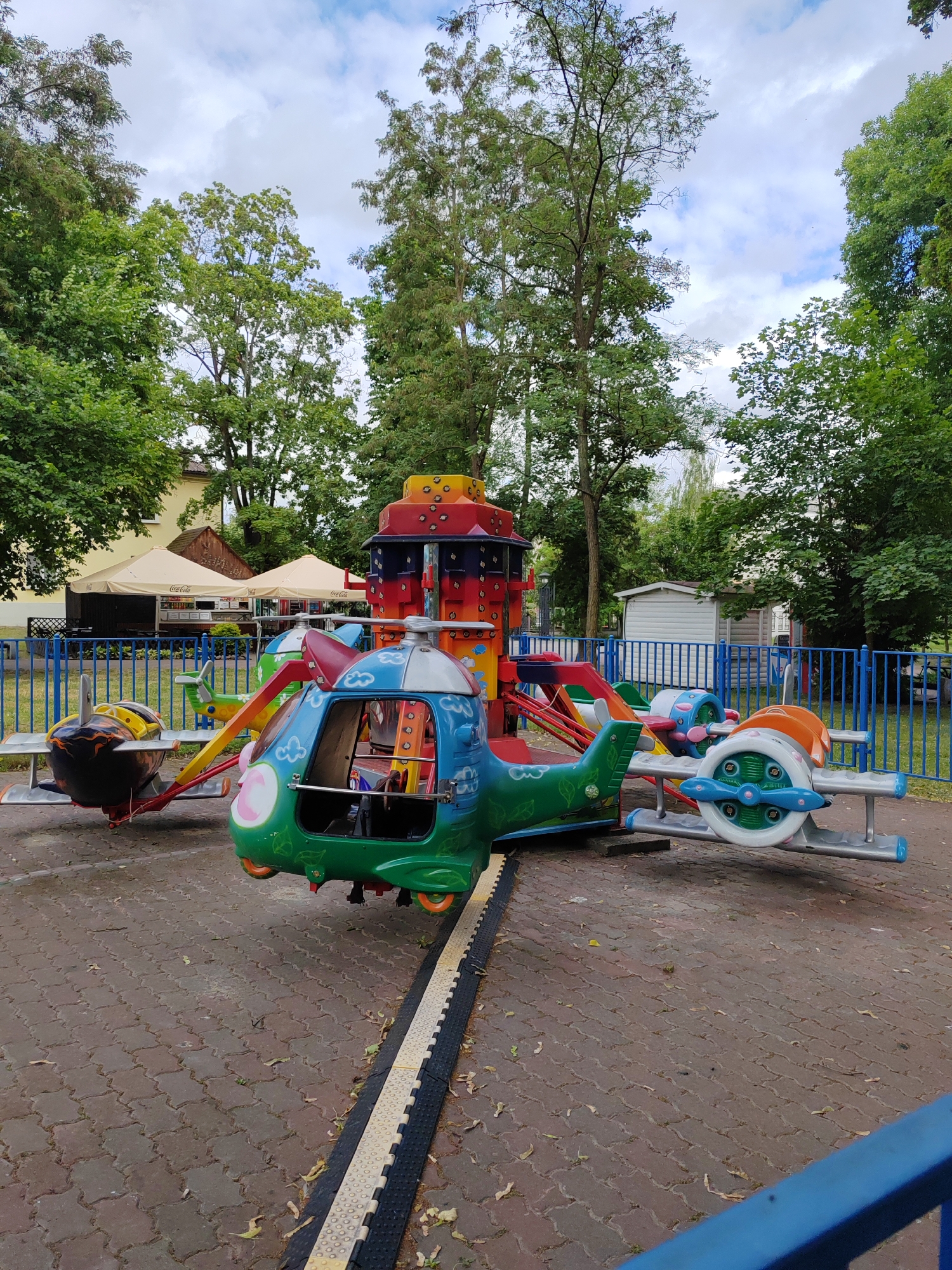 The image depicts a children's amusement ride with various aircraft-themed vehicles, including a helicopter and an airplane, attached to a central rotating structure. The ride is set up on a paved area with a brick-like pattern. In the background, there are trees, a building with awnings, and a blue metal fence. The sky is partly cloudy. This scene is located in a park or amusement area within the city of Brest, Belarus. There are no people visible in the image.