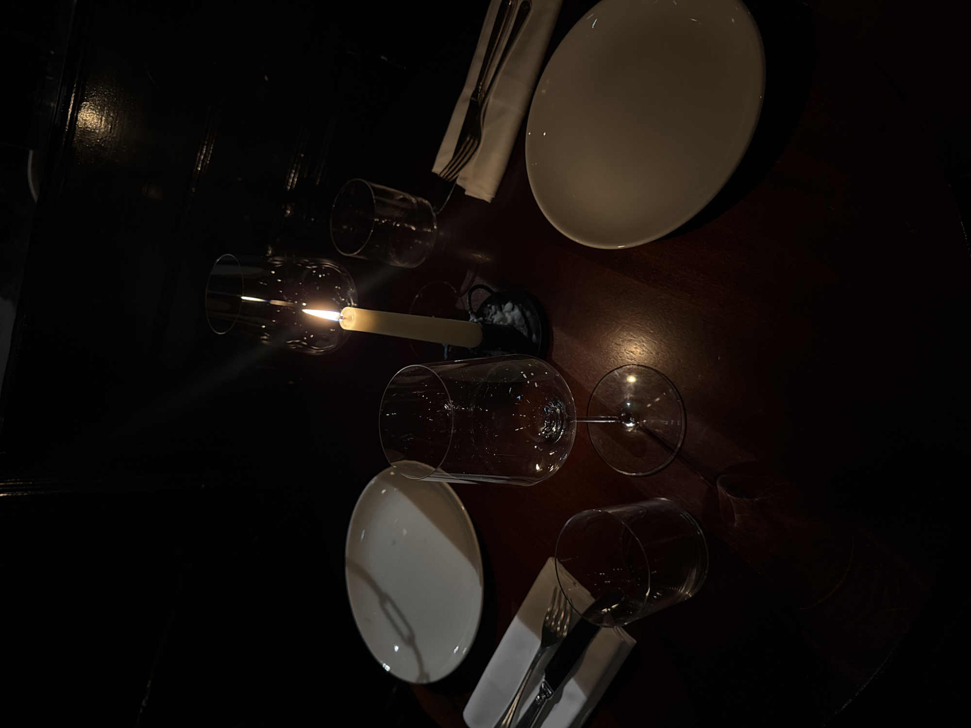 This image depicts a dimly lit indoor dining scene, likely a restaurant or similar establishment, located in the city of London, United Kingdom. The foreground features a dark, polished wooden table, partially illuminated by a single, cream-colored candle burning in a dark, possibly metallic, holder near the center-right, with visible wax drippings. The table is set for two people, though no individuals are present.
On the left side of the table, an empty, round white dinner plate is positioned. To its left, a white folded napkin holds a silver fork and knife. A clear, empty drinking glass is situated in front of the napkin. A tall, empty stemmed wine glass is placed slightly to the right of this setting.
On the right side of the table, another empty, round white dinner plate is visible. To its upper right, a white folded napkin contains a silver fork and knife. An additional clear, empty drinking glass is positioned above this napkin. A second tall, empty stemmed wine glass is visible further back, near the candle. The background is dark and indistinct, suggesting an interior wall or paneling. There are no observable actions or interactions occurring.